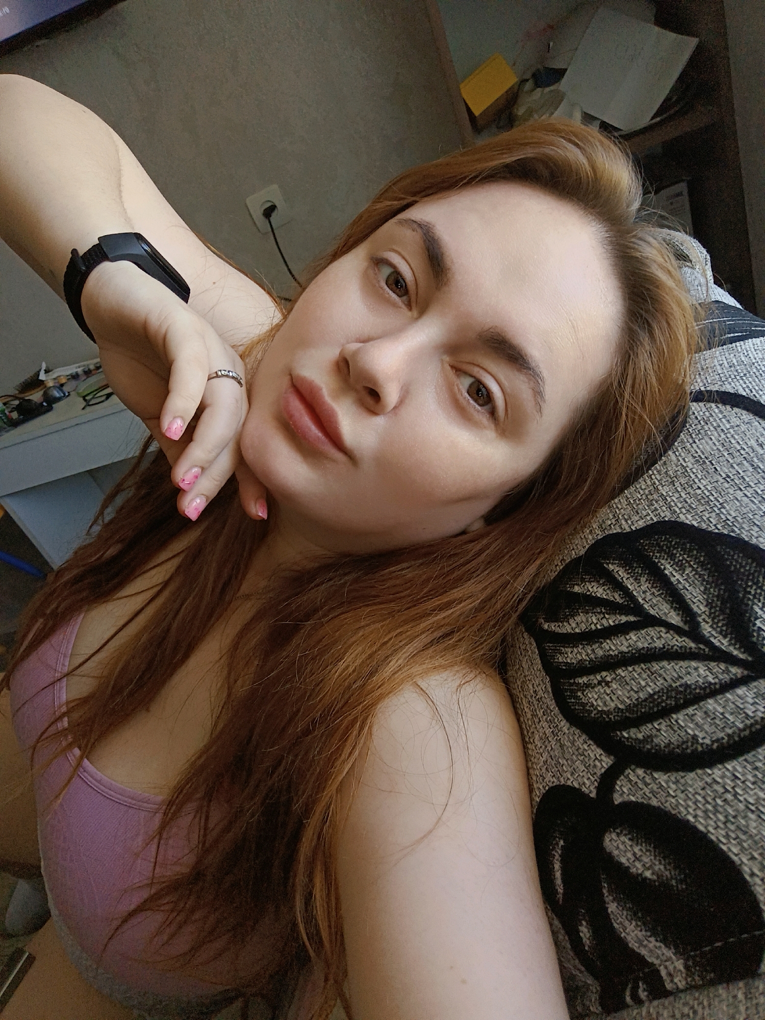 A young, fair-skinned woman with reddish-brown hair is reclined, possibly on a sofa or bed, with a gray and black patterned pillow behind her head. She is wearing a light pink tank top. Her left arm is raised, with her hand near her face; she has a black fitness tracker on her wrist and a ring on her finger. Her nails are painted light pink. The background includes a desk with various items, an electrical outlet, and furniture with objects stacked on top. The location is potentially indoors in Chaikovsky, Russia, given the prompt's specification.