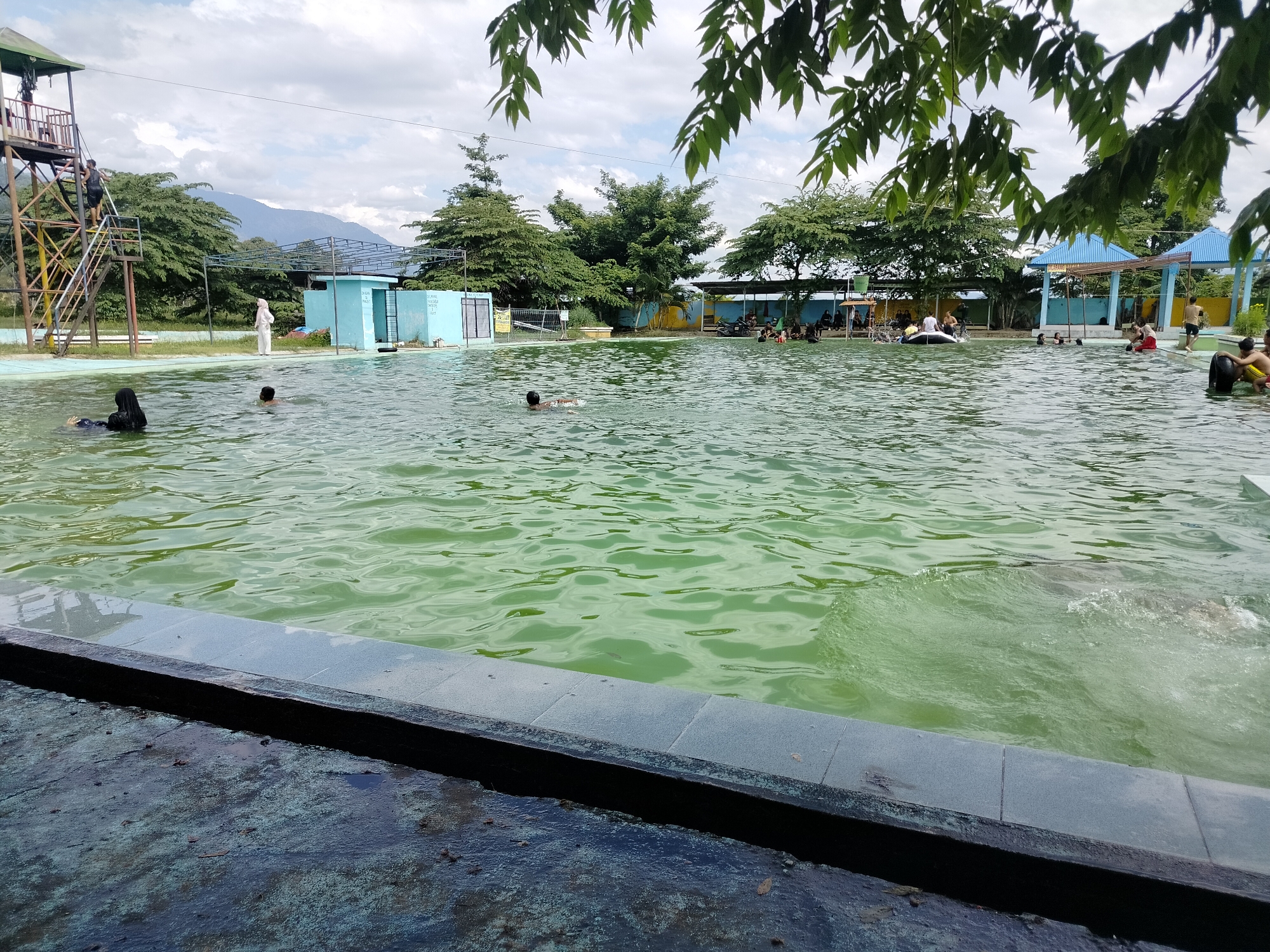 The image shows an outdoor swimming pool in Tegarmiko, Indonesia. The pool water is a murky green color. Several people are swimming and playing in the pool. On the left side of the pool, there is a tall, metal observation tower with stairs. In the background, there are trees, buildings, and mountains. The sky is cloudy.