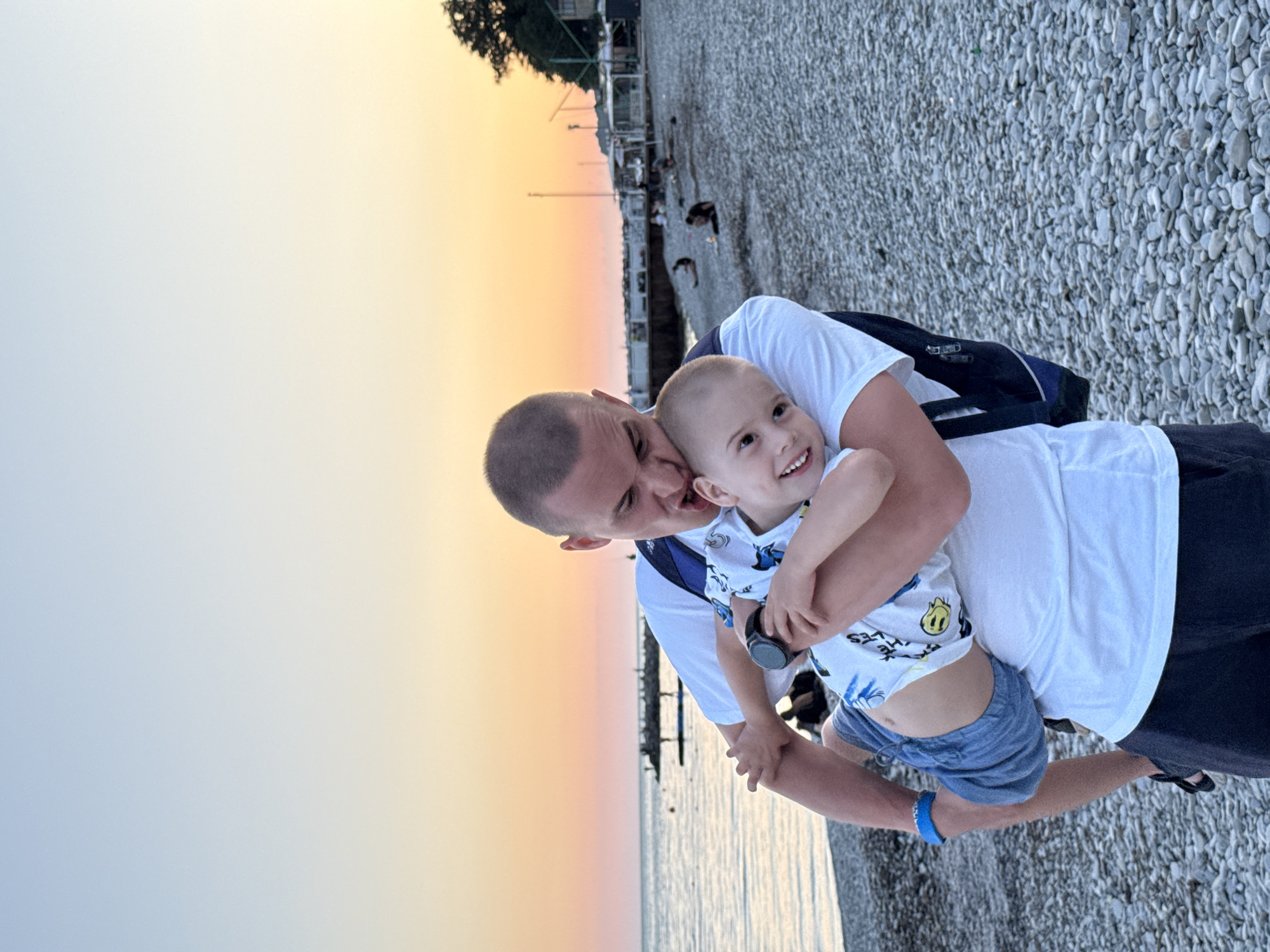 The image shows a man and a young child on a pebble beach, likely near the sea, given the water visible in the background. The sky suggests it is either sunrise or sunset, with warm hues of orange and yellow transitioning to a pale blue. The man, wearing a white t-shirt and a backpack, is holding the child, who is also in a white t-shirt and blue shorts. The child is smiling widely, looking upwards, and seems to be enjoying the embrace. The man is leaning in towards the child, appearing affectionate. Behind them, the beach is covered in small grey pebbles, and a few other people are visible in the distance, along with some structures that might be part of a pier or promenade. The overall mood is warm and joyful, capturing a tender moment between the two individuals. The child is wearing a blue bracelet on his left wrist. Some faint text is visible on the child's shirt, appearing to be part of a design.