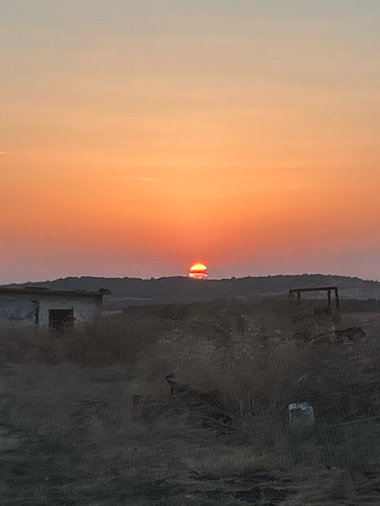 The image depicts a sunset in Krepost, Bulgaria. The sun, partially obscured by a rolling hill, casts a warm orange glow across the sky. In the foreground, dry, overgrown vegetation covers the ground. To the left, a dilapidated, concrete structure with an open doorway stands abandoned. In the middle ground, a rectangular metal frame, possibly part of a forgotten structure, is visible. The overall scene conveys a sense of desolation and the quiet end of a day.
