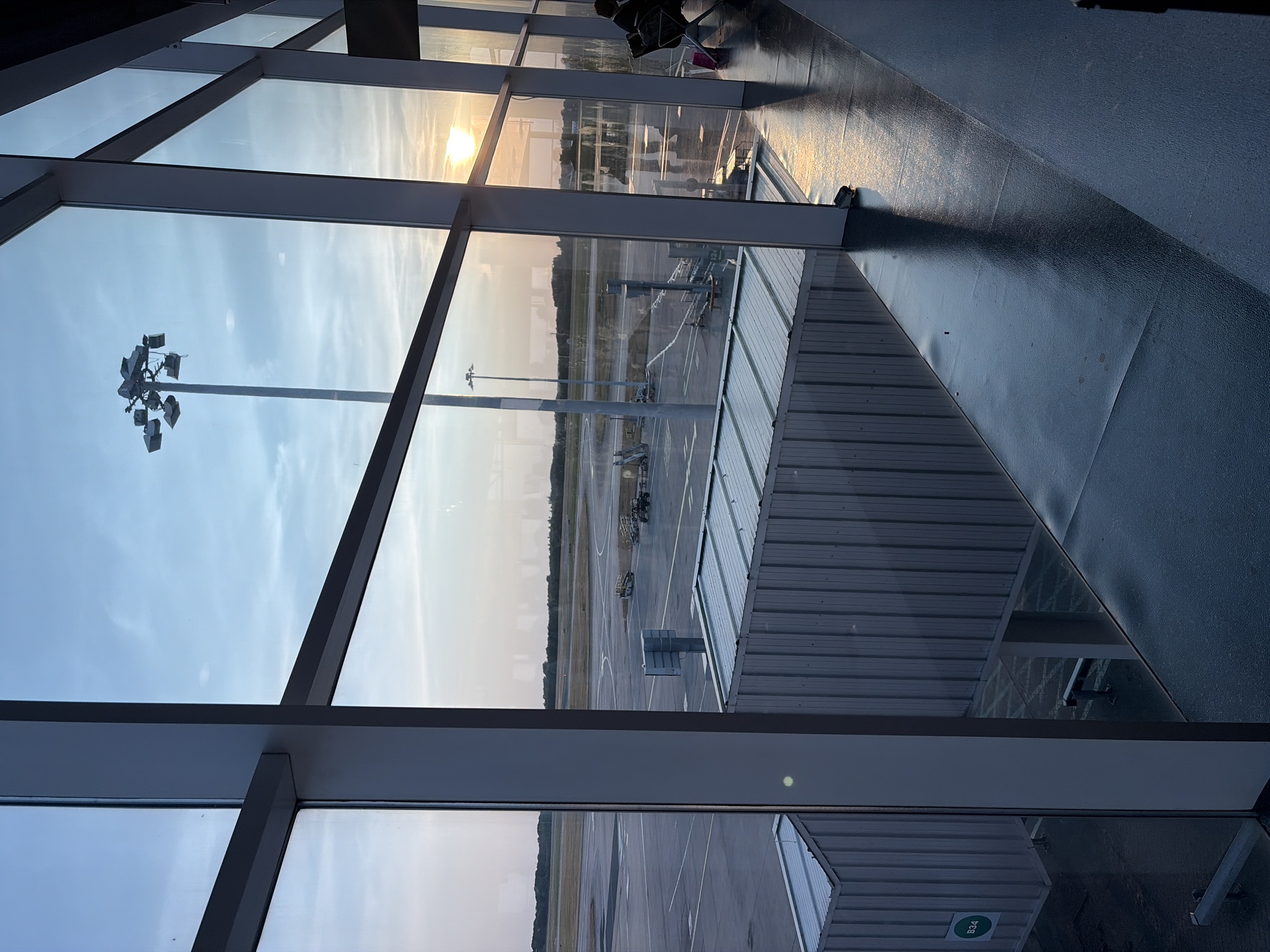This media file depicts an airport scene viewed from an interior, likely a departure lounge, looking out through a large, multi-paned window. The scene is set at an airport, with runways, taxiways, and various airport equipment visible. A jet bridge extends from the building towards an unseen aircraft. The time of day appears to be either early morning or late afternoon, given the soft, golden light of the sun setting or rising, casting reflections on the glossy floor of the interior. The sky is partly cloudy.
In the foreground, the interior of the airport is visible, characterized by large glass windows and dark, reflective flooring. The windows are framed by dark metal beams, creating a grid-like structure. Chairs or lounge seating are partially visible in the upper right corner, suggesting people are present or have recently been there, though no individuals are clearly depicted. A tall light fixture with multiple lamps stands prominently on the tarmac, a common feature of airport exteriors.
The exterior scene shows a typical airport tarmac with paved surfaces, markings, and several vehicles or pieces of ground equipment. Trees line the horizon in the distance, indicating a rural or semi-rural location. The jet bridge has a ribbed, metallic appearance. A circular green sticker with the white text "B34" is visible on a white structure near the bottom right of the image, likely indicating a gate or boarding area number. The overall mood is one of quiet observation, perhaps anticipation or departure, with the warm light suggesting a peaceful transition. The location is identified as Uttlesford, United Kingdom.