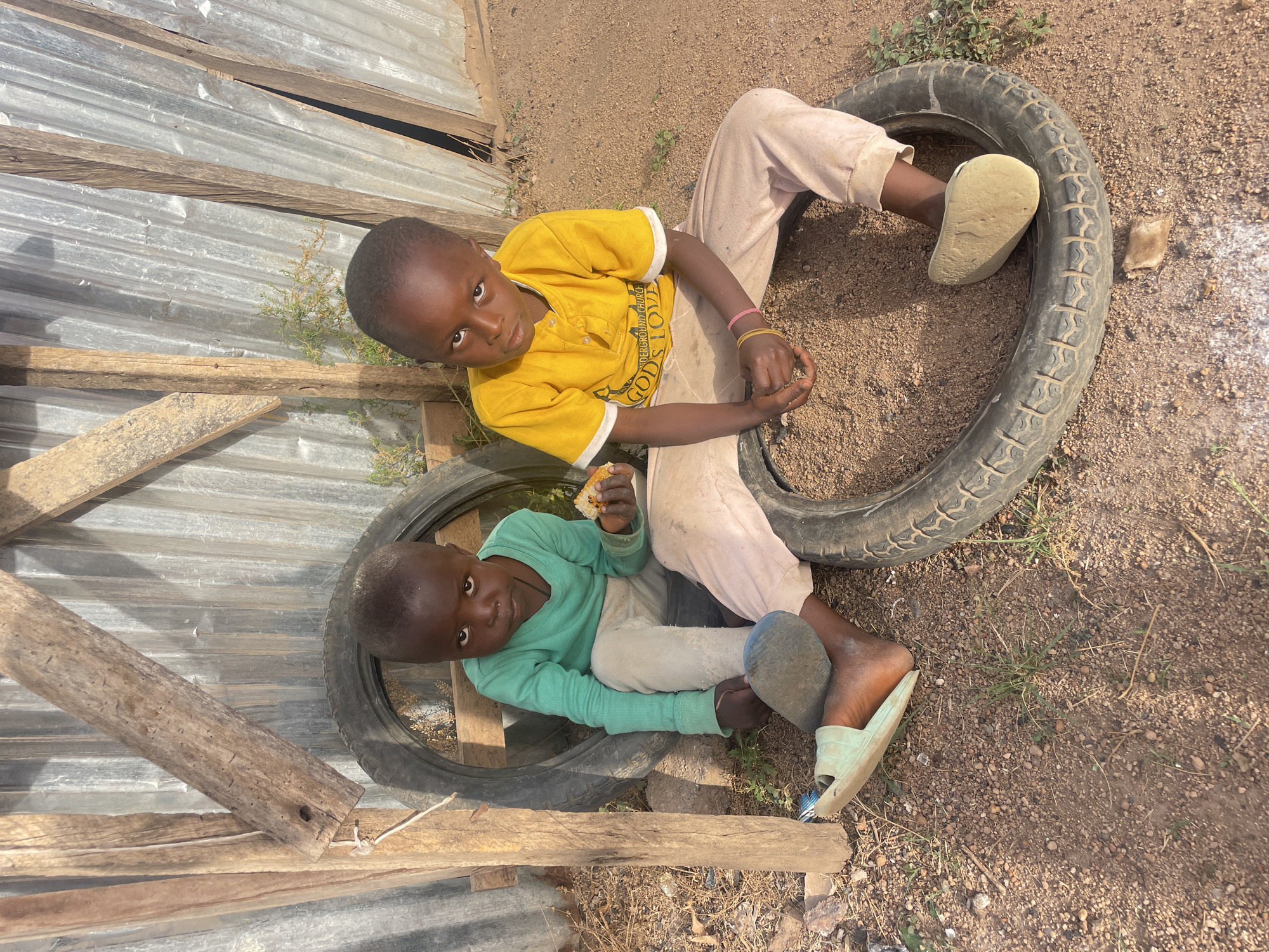 This is a top-down photograph depicting two young boys sitting on old vehicle tires outdoors in Barakin Lerawi, Nigeria. The ground is dry, dusty earth mixed with small pebbles and sparse patches of dry grass, suggesting a sunny and possibly arid environment. To the left and in the background, a rudimentary structure or fence constructed from corrugated metal sheets and rough wooden planks is visible.
The boy in the upper portion of the frame wears a bright yellow short-sleeved shirt with white cuffs and light-colored long pants. He has short dark hair and a dark complexion, looking directly at the camera with a neutral expression. His yellow shirt bears partially visible English text, reading "UNDERGROUND CHURCH" and "GOD'S LOVE" (or "GOD'S TOVE"). He has pink and red elastic bands on his left wrist.
The second boy, situated lower and slightly to the left, is dressed in a light green long-sleeved shirt and light-colored long pants, one of which has a tear near the knee. He also possesses short dark hair and a dark complexion, gazing directly at the camera. He holds a piece of bread or pastry in his right hand, appearing to be eating it. His left foot is partially covered by a light blue or green slide sandal.
Both children are using the tires as seats. The bright, clear lighting indicates it is daytime. The scene portrays a moment of daily life in a simple, outdoor setting.