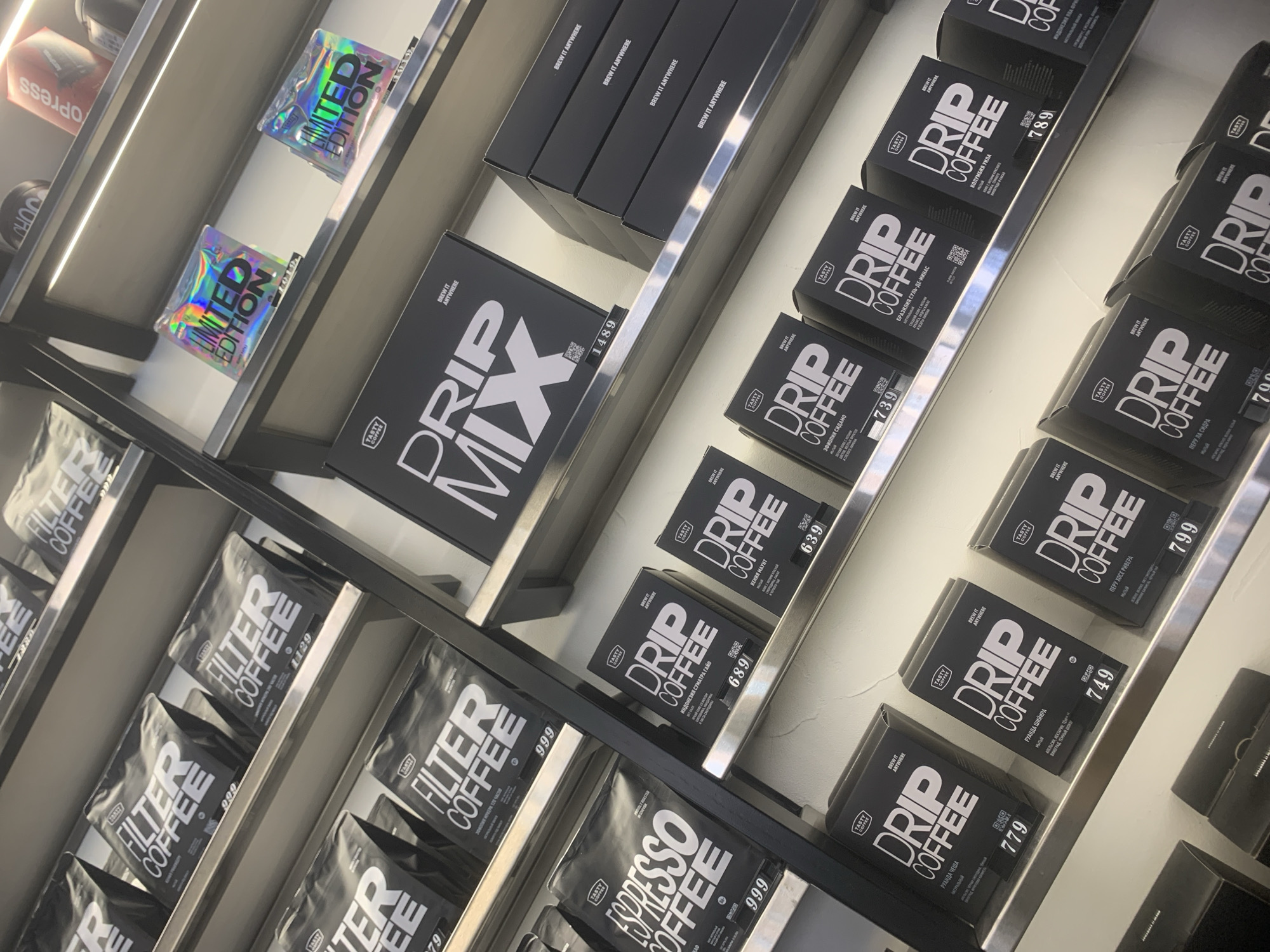 The image shows a product display on a shelf in a store, possibly in Izhevsk, Russia, based on the OCR text. The products include various types of coffee, such as "Drip Coffee", "Filter Coffee", and "Espresso Coffee," packaged in black containers and bags. There are also black boxes and colorful "Limited Edition" foil packs. The OCR text includes information about the origin of the coffee beans (e.g., "Indonesia Sumatra Gayo," "Ethiopia Sidamo") and the flavor profiles (e.g., "Orange, Nectarine, Tea", "Grapes, Dark Chocolate"). There are also monetary values listed on some of the products. The shelves are silver-colored with built-in lighting.