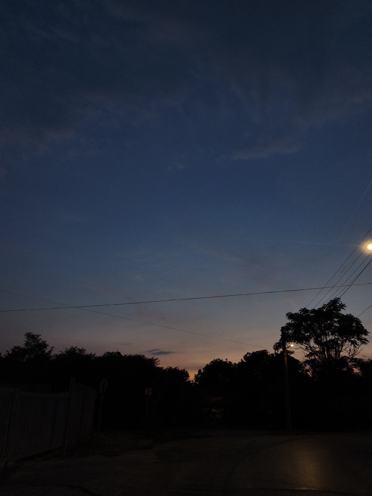 The image depicts a rural road at dusk, with a gradient of blues and oranges in the sky. Silhouetted trees form a dark line along the horizon. A street light illuminates the area, casting a warm glow on the road ahead. In the foreground, a fence runs along the left side of the road. The scene appears tranquil and serene. The location is identified as Lovech, Bulgaria.