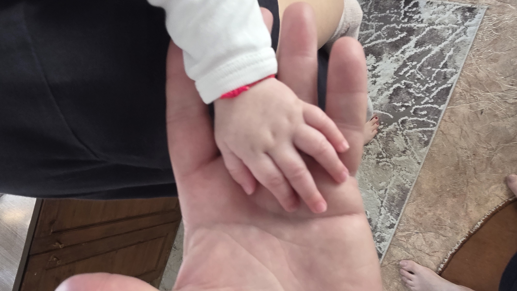 A close-up shot reveals a baby's hand, wearing a white sleeve with a red band at the wrist, resting on a larger adult hand. The adult is wearing dark pants. In the background, a portion of a rug with a gray and white marble pattern sits next to a tan-colored floor. Fragments of wooden furniture and feet are visible at the periphery. There's nothing in this image that indicates this was taken in Blagoevgrad, Bulgaria.