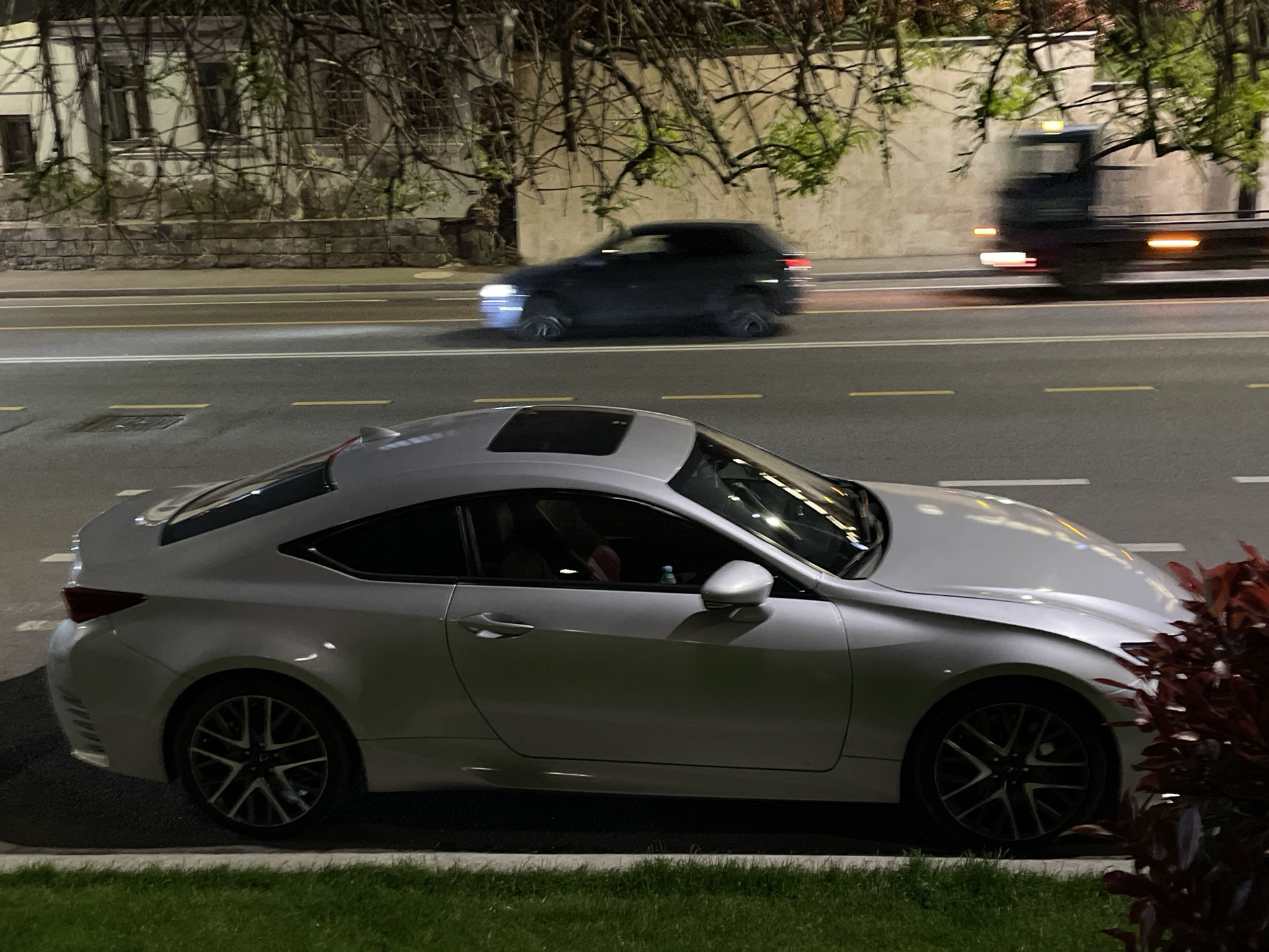The image captures a nocturnal street scene in Tbilisi, Georgia, with a prominent light-colored (likely silver) Lexus RC coupe parked in the foreground. The car, featuring a sunroof and intricate multi-spoke wheels, is situated on a grassy verge beside a paved sidewalk.
Beyond the parked vehicle, a multi-lane asphalt road stretches horizontally across the frame, marked with clear white solid lines and broken yellow lines. Two vehicles are in motion on the road, depicted with significant motion blur, indicating they are moving at speed. One is a dark SUV or crossover, visible primarily by its bright headlights. Further in the background, a large, dark truck is also in motion, its tail lights and side markers appearing as streaks of red and orange light.
The background features a building with multiple windows and a stone foundation, partially obscured by a network of bare tree branches with some sparse green foliage. The scene is illuminated by artificial streetlights, casting ambient light and creating reflections on the parked car's surface. The overall impression is a quiet street at night with some passing traffic. There is no visible text.