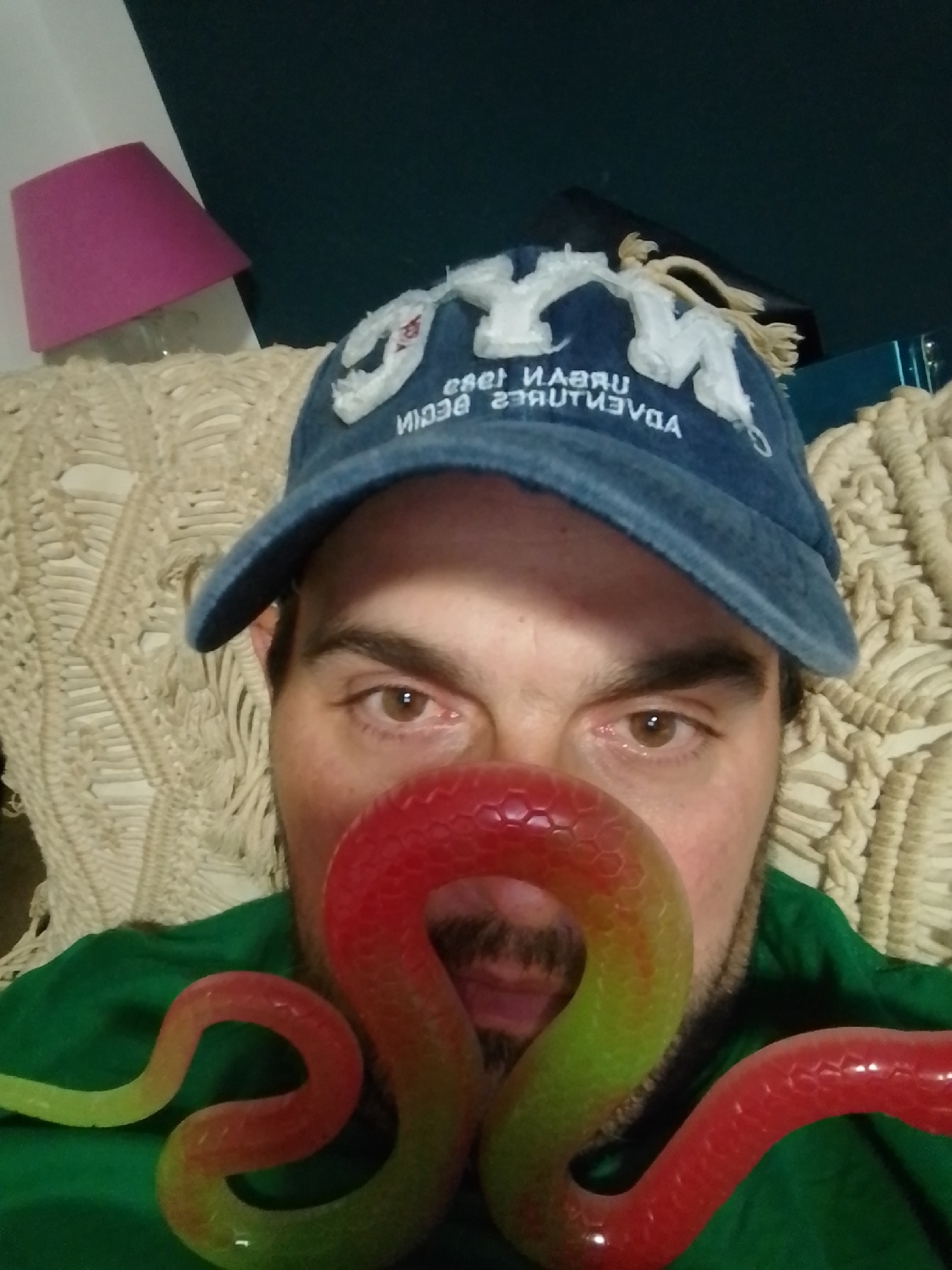 A man wearing a blue baseball cap and a green shirt is photographed from the chest up. The cap has white lettering that appears to be "URBAN ADVENTURES" and some other obscured text. A colorful, snake-like object, transitioning from red to green, is positioned in front of the man's face, obscuring his nose and mouth. The background consists of a textured, cream-colored fabric, possibly a sofa or blanket, and a pink lampshade is visible in the upper left corner. The scene appears to be indoors, in a domestic setting. There is no discernible connection to the city of Wedel, Germany.