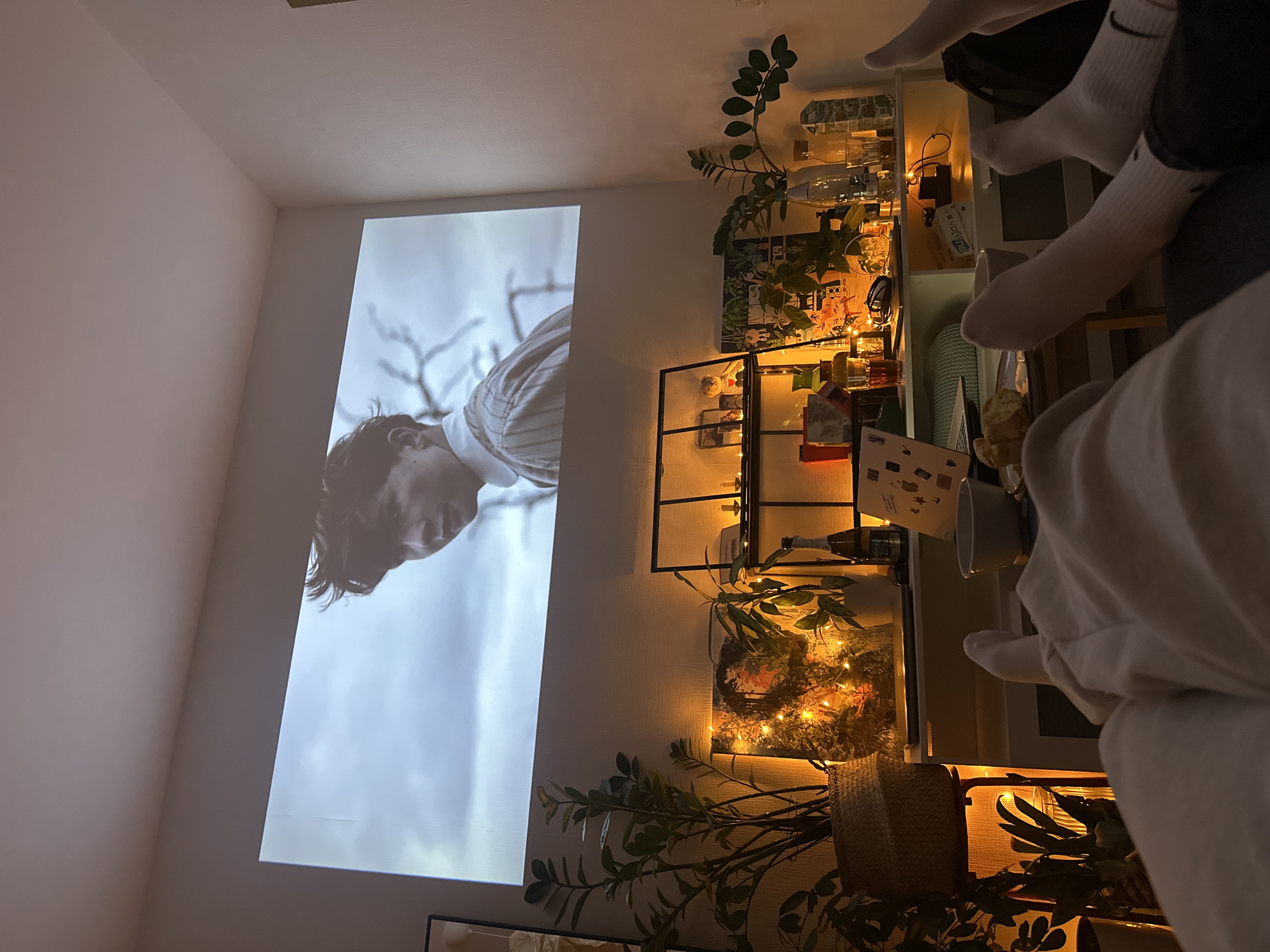 A scene indoors showing a projected image on a white wall, viewed from a slightly tilted perspective. The projection displays a man looking upwards against a sky backdrop. To the right of the projection, a shelf unit is adorned with plants, glassware, and decorative lights. In the foreground, legs clad in white socks extend towards the camera. The overall setting suggests a cozy, domestic environment, possibly in an urban apartment.