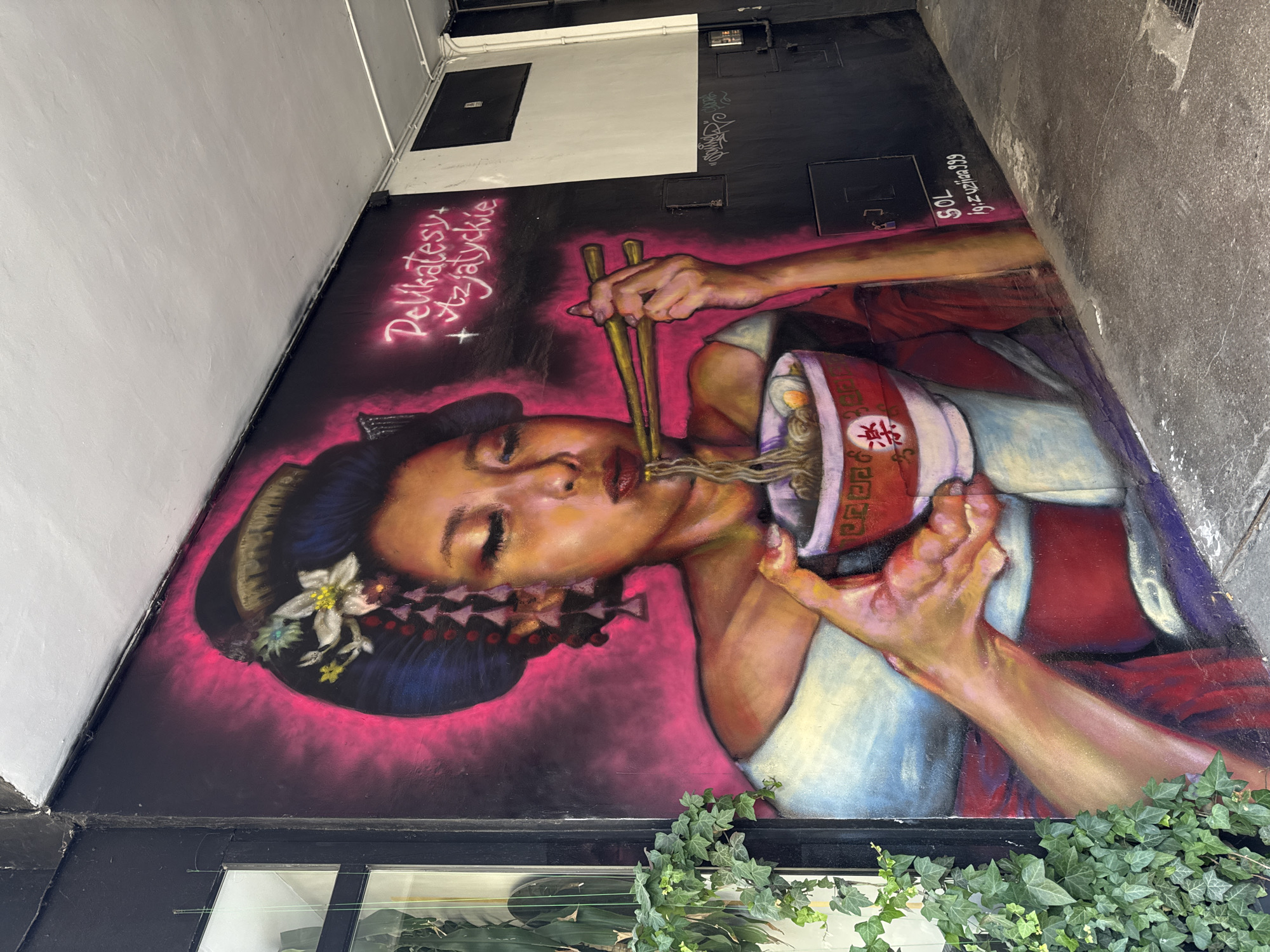 The image depicts a mural on a building exterior. The mural features a woman in traditional Japanese attire, with her hair styled in an updo and adorned with flowers and ornaments. She is depicted with her eyes closed, holding chopsticks and eating from a bowl of ramen noodles. The mural is vibrant, with a hot pink background that fades to black. Text in neon pink reads "Delikatesy + Azjatyckie," which translates to "Delicatessen + Asian." Below this, on the lower right side of the mural, are two lines of text in white spray paint: "SOL" and "ig: z uztina_999". The mural is located on a narrow alleyway in Poznan, Poland, as indicated by the Polish text on the mural. A modern, dark-colored metal door frame and a glass pane are visible to the left of the mural, with green ivy plants growing alongside. The ground in front of the mural is a rough, grey concrete surface. The overall scene suggests an urban setting, possibly a street art installation in a commercial or pedestrian area.