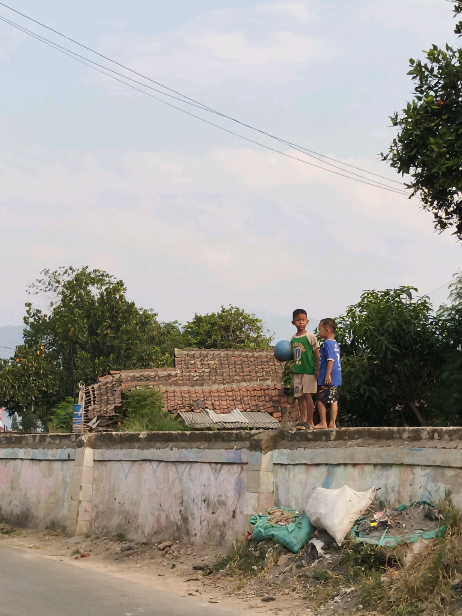Two young boys stand on top of a low, decorated concrete wall in Panyadap, Indonesia. The boy on the left wears a green t-shirt and light-colored shorts, holding a blue ball. The boy on the right wears a blue t-shirt and dark patterned shorts. Behind them, a cluster of tiled roofs of a building is visible, partially obscured by lush green trees. The sky above is a pale blue with some cloud cover. To the right of the boys, a large green tree with dark leaves is prominent. Below the wall, the ground is littered with trash, including two green bags and a large white bag filled with debris. A paved road runs along the bottom of the image. The scene appears to be a residential area.