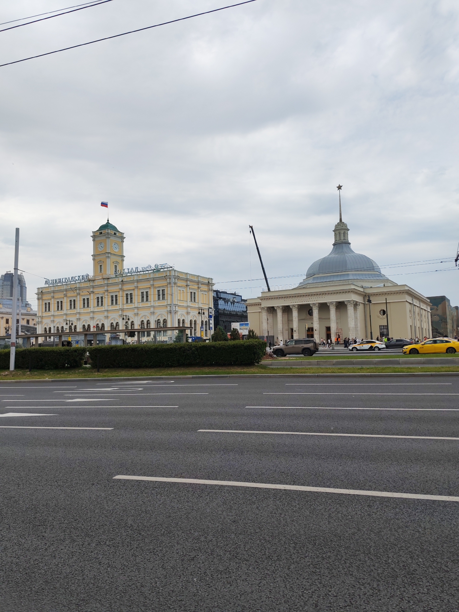 The image depicts a view of Leningradsky railway station in Moscow, Russia, from a street. In the foreground, a grey asphalt road with white lane markings is visible. Beyond the road, a manicured hedge separates the street from a walkway with a glass barrier.
The main subjects are two prominent buildings. The building on the left is the Leningradsky railway station itself, a light yellow structure with a prominent clock tower topped by a green roof and a Russian flag. The building features arched windows and a sign that reads "ЛЕНИНГРАДСКИЙ" (Leningradsky) and "ВОКЗАЛ" (Vokzal). The building on the right is a larger, domed structure with classical columns and a spire topped with a red star. People are visible walking in front of this building. Several cars, including yellow taxis and a dark SUV, are parked or driving on the road in front of the buildings. A construction crane is positioned behind the domed building. The sky is overcast with grey clouds.