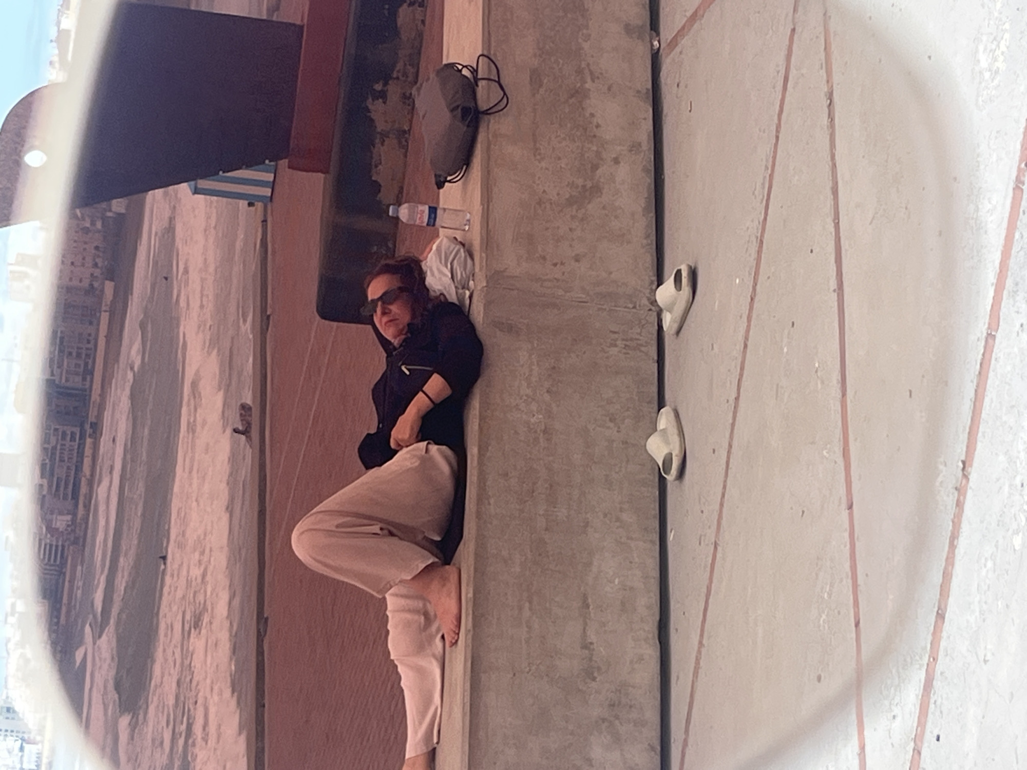 The image captures a woman resting outdoors on a concrete surface, likely a rooftop, terrace, or balcony, under bright daylight. She is lying on her back, wearing sunglasses, a black long-sleeved top, and light beige or pinkish-hued pants. Her bare feet are visible, suggesting a relaxed posture.
Adjacent to her head on the concrete surface are a grey backpack or duffel bag, a white folded garment (possibly a t-shirt), and a clear plastic bottle of Evian water. Further away from her feet, two white open-toed sandals or slippers are placed on the concrete ground.
The background on the left side of the image is seen through a circular, semi-transparent effect, possibly a reflection in her sunglasses or a window. This view reveals a distant cityscape featuring numerous buildings, and a wide sandy area or beach, consistent with a coastal urban environment like Las Palmas De Gran Canaria. A dark, broad structure partially overhangs this background view, with a blue and white striped object visible beneath it. The overall atmosphere conveys relaxation in a sunny, urban-coastal setting.