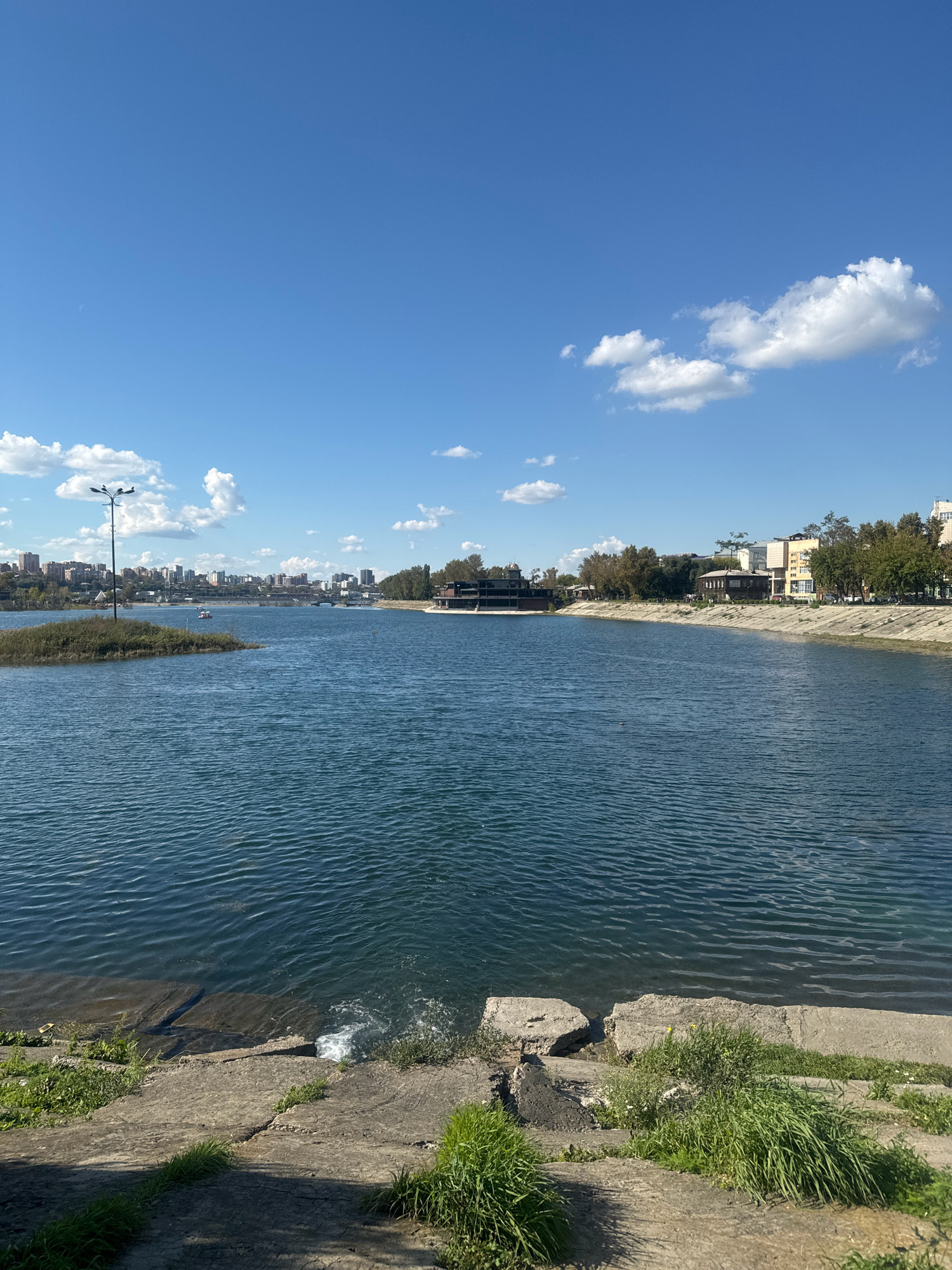 The image depicts a wide body of blue water, likely a river or lake, under a clear blue sky with scattered white clouds. In the foreground, a concrete embankment with some patches of green grass and sparse vegetation slopes down to the water's edge. The water exhibits ripples, suggesting a gentle breeze.
Across the water, the opposite bank is lined with trees and buildings, indicating an urban environment. A prominent structure resembling a restaurant or cafe with a dark roof sits near the water on the right side of the image. Further down the shore, buildings of varying heights and architectural styles are visible, interspersed with more trees.
On the left side of the water, a small island covered in reeds is present, with a lamppost on top. In the distance, a cityscape with numerous buildings forms the horizon, and a bridge can be seen spanning the water. Several small boats are scattered on the water, with one boat appearing to be a paddle boat with people on it.
Based on the architectural style of the buildings and the presence of the Angara River, this scene is likely located in Irkutsk, Russia, specifically along the embankment of the Angara River. The overall atmosphere is one of a pleasant day in a city situated near a substantial body of water.