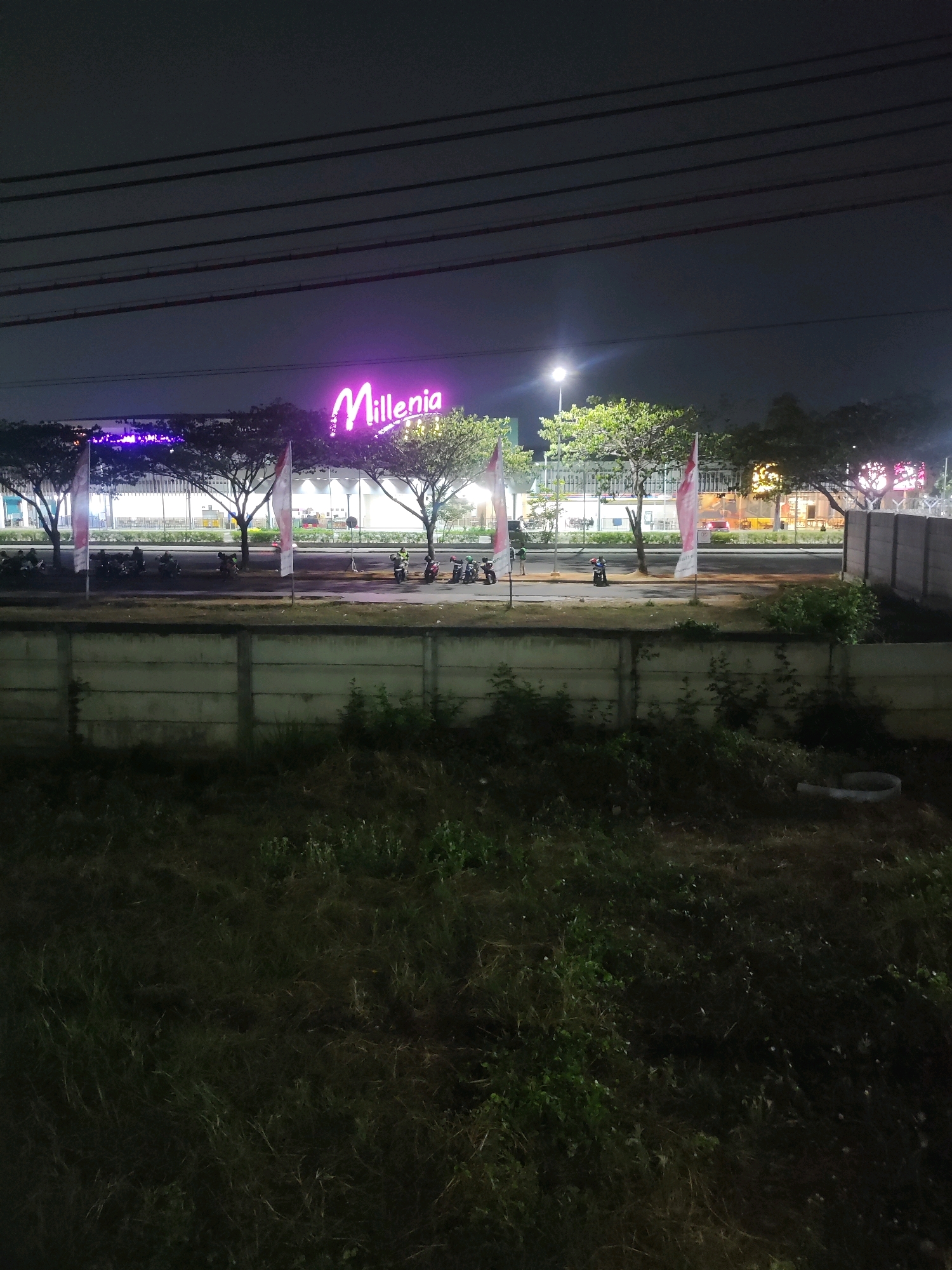 An eye-level, medium shot captures a nighttime view of the "Millenia" building in Telagamurni, Indonesia. The building is illuminated with vibrant purple neon lights forming its signage. In front of the building, trees and flags are visible, with a row of motorcycles parked on the road. A concrete wall separates the road from a grassy area in the foreground. The sky above is dark, with visible power lines.