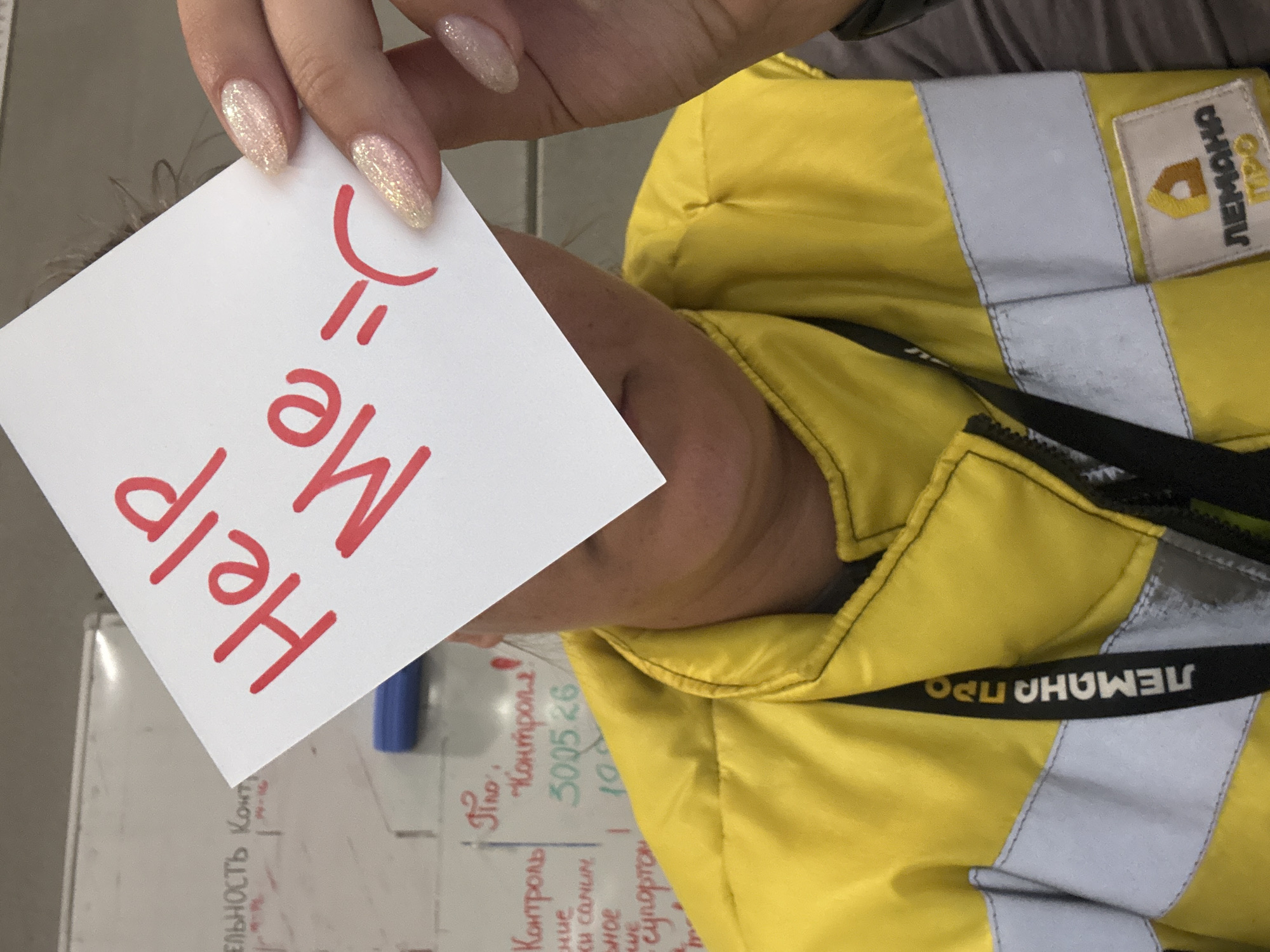 This close-up image, likely taken in Lupanovo, Russia, captures a person wearing a bright yellow work or safety jacket featuring reflective strips and black straps. The jacket has visible branding with the text "ЛЕМАНА ПРО" (LEMANA PRO) in Russian on both a tag and a strap, consistent with the provided location context.
A hand with manicured, glittery light-colored nails holds a white piece of paper. Written on the paper in red marker, and oriented upside down relative to the viewer, are the words "Help Me =(". This handwritten note, accompanied by a sad/tired emoticon, suggests a humorous or lighthearted expression of being overwhelmed, rather than an emergency.
In the blurred background, a whiteboard is visible, filled with handwritten Russian text and numbers. Phrases like "Контроль" (Control), "Типо" (Type), and numerical entries such as "3005 26" and "100" can be deciphered, indicating a work-related setting like an office, warehouse, or a control room. A blue marker also lies near the whiteboard. The indoor lighting suggests a typical daytime work environment.