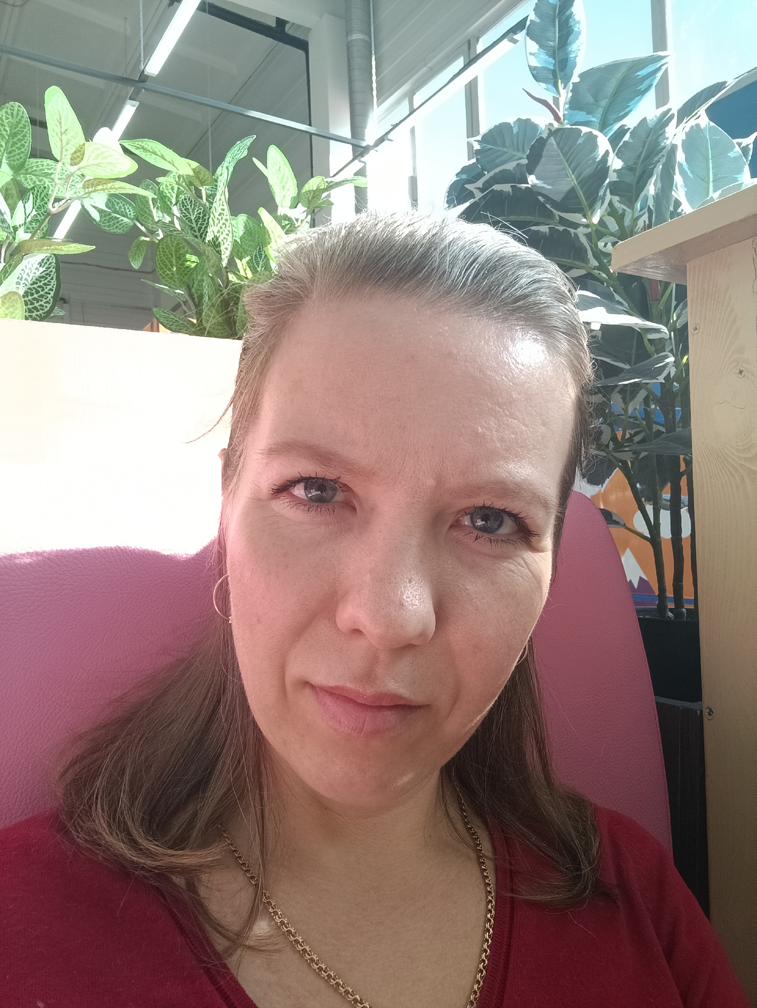 A woman with fair skin and blue eyes is facing the camera. She has blonde hair, styled back, and is wearing a red v-neck shirt and a gold chain necklace. A small gold hoop earring is visible in her left ear. She is seated in a pink chair. Behind her, there are two potted plants, one with green and white patterned leaves and the other with large green and white leaves. The background includes a window with a view of a bright sky. The interior setting appears to be a room with industrial-style lighting fixtures and a wooden structure. The exact location in the city of Chaikovsky, Russia, cannot be determined from the image.