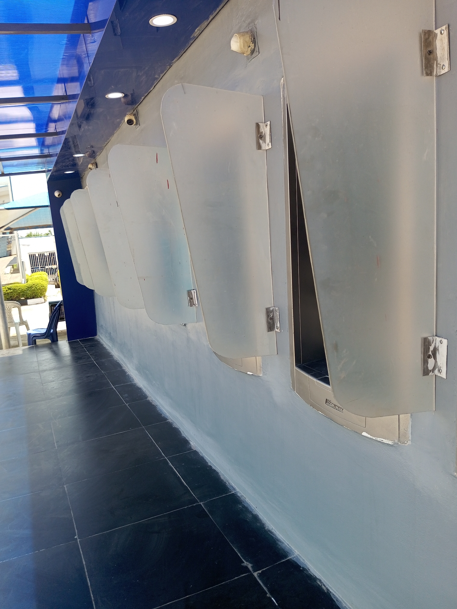 A row of seven stainless steel urinals with frosted glass dividers is mounted on a light gray concrete wall in Bukuru, Nigeria. The floor is tiled with large black squares. A blue corrugated plastic roof extends over the urinals, supported by metal beams. Lights are embedded in the blue ceiling. Two small security cameras are visible above the urinals. In the background, several white plastic chairs and a blue plastic chair are visible next to a building with a barred window and lush green bushes. The scene appears to be an outdoor or semi-outdoor restroom facility.