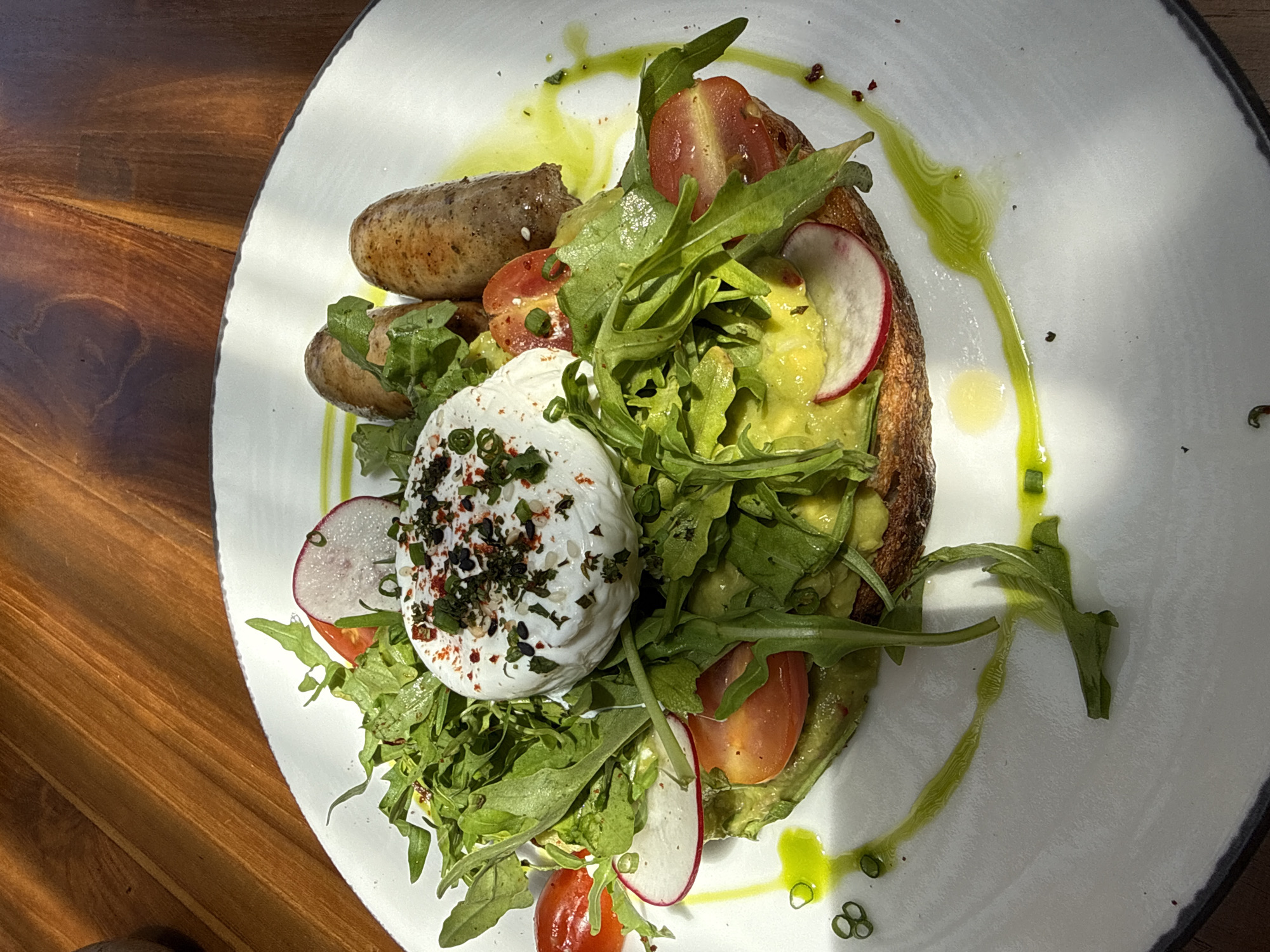 A top-down, medium shot depicts a white plate holding a composed breakfast dish. The dish features avocado toast with sliced tomatoes, radish, and a poached egg topped with seasonings. Arugula is liberally used as a garnish. Two sausages rest to the left of the toast. A drizzle of green sauce, possibly herb oil, is decoratively swirled around the plate. The plate is set on a wooden table. The location of Kuta Selatan, Indonesia is not discernible from the image alone.
