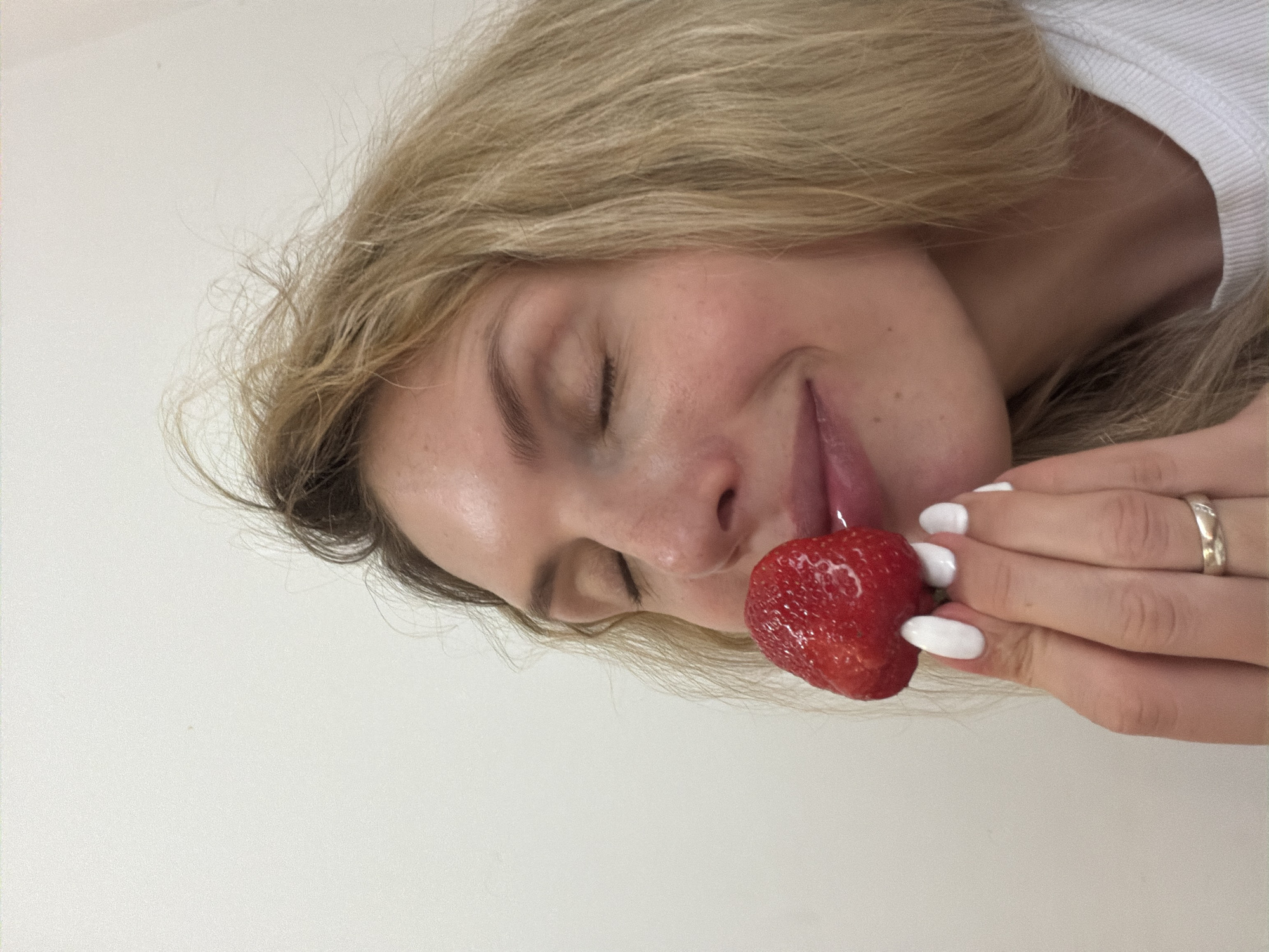 A woman with blonde hair and closed eyes is holding a red strawberry up to her lips, with her tongue slightly protruding. She is wearing a white shirt, and a silver ring is visible on her finger. The background is a plain white surface. The location in the city of Budva, Montenegro is not discernible from the image.