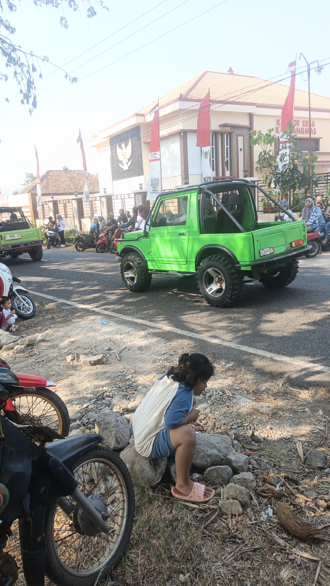 A young girl with dark hair, wearing a blue and white t-shirt and blue shorts, sits on rocks by the roadside in Kandangmas, Indonesia. She is wearing pink slides. In the background, a bright green open-top vehicle is parked on the street, with people visible inside and around it. Several motorcycles are also present, some parked and others with riders. A building with Indonesian flags is visible behind the vehicles. The ground is covered with gravel, dry grass, and scattered leaves. The scene appears to be outdoors during daylight hours.