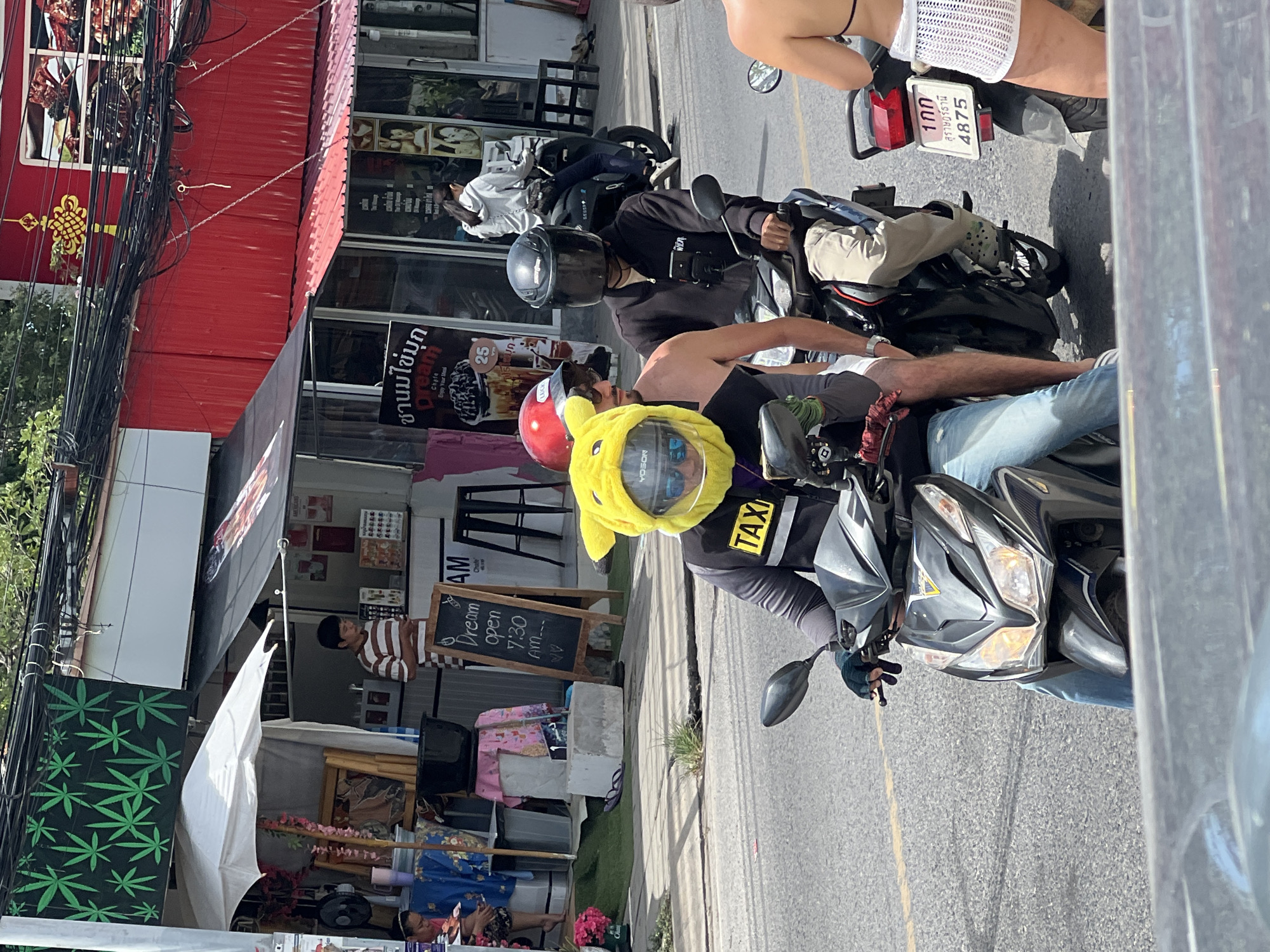The image shows a street scene in Baan Chaweng, Thailand. Three motorbikes are on the road. The foremost rider wears a yellow Pikachu helmet with a "TAXI" emblem on their vest. The passenger behind them wears a red helmet. Behind them, another person on a motorbike wears a black helmet. Further back, another rider wears a white crocheted skirt. A sign reads "Dream open 7:30 AM". A person is visible in a striped shirt near the store. The word "Dream" is visible on another sign, with Thai script. A green leaf print is displayed near the sidewalk.