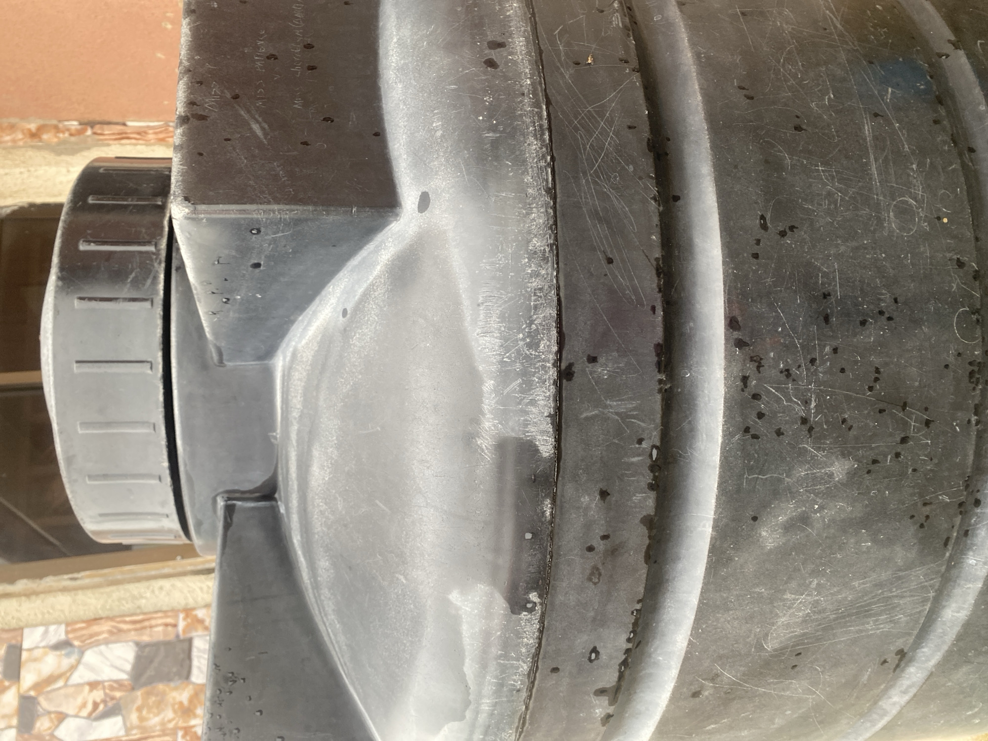 The image shows a close-up of a large, black, plastic water tank. The tank has a ribbed texture and appears to be weathered, with numerous scratches and some dark marks on its surface. A silver, ribbed lid is visible on top of the tank. The background shows parts of a building with textured walls and what appears to be a window or opening. There are some faint inscriptions on the tank's surface, including "MISS P", "MISS PATIENCE", and "MISS LIBERAL (ANGA)". The scene is consistent with an outdoor setting in Jauro Yinu, Nigeria, where such water tanks are commonly used for water storage.