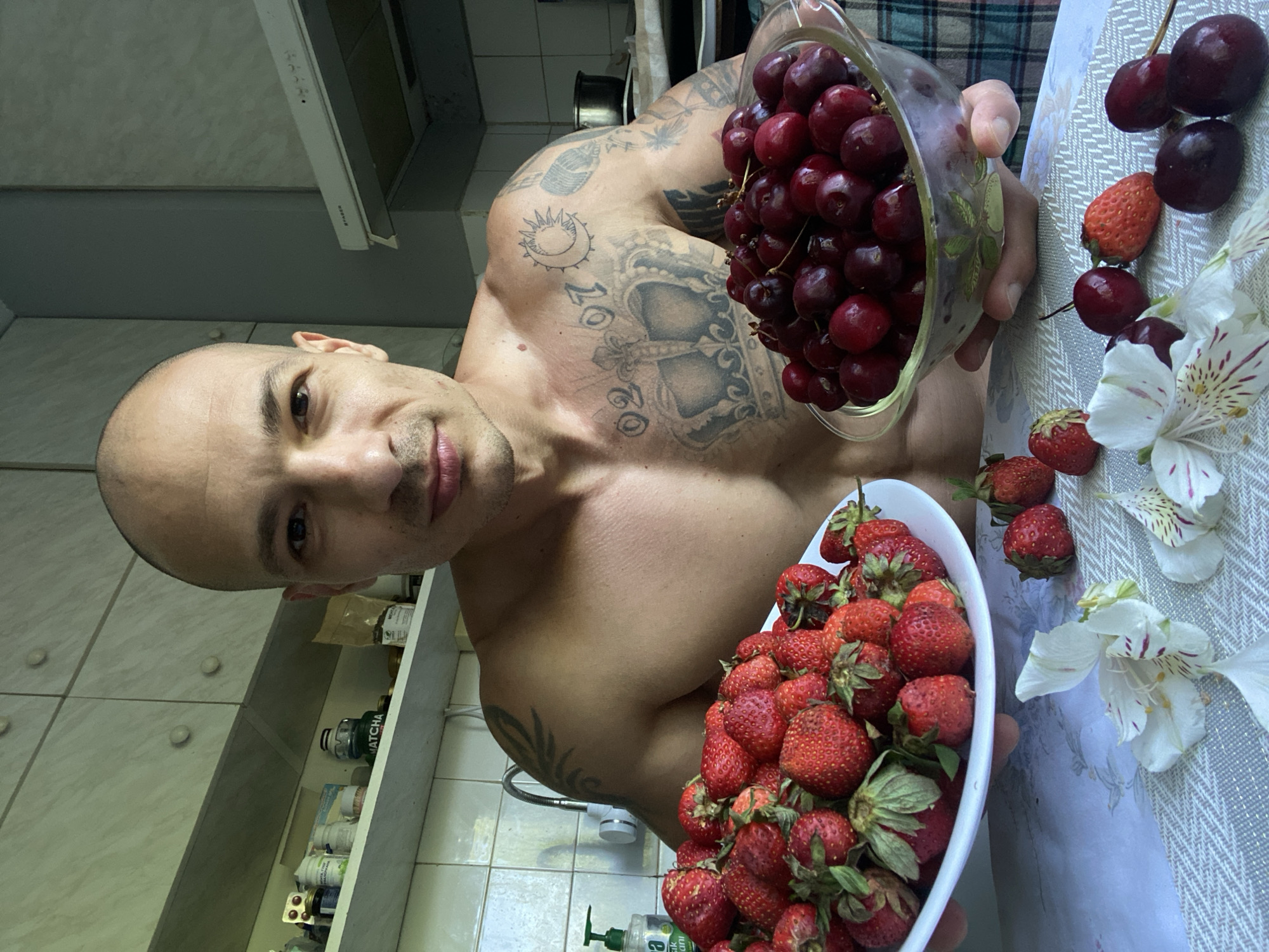A shirtless bald man with extensive tattoos on his chest and arms is in a kitchen holding two bowls of fruit. In his left hand, he holds a white plate piled high with fresh strawberries. In his right hand, he holds a clear glass bowl filled with dark red cherries. The kitchen appears to be in an apartment in Pleven, Bulgaria, with light-colored cabinets and tiled walls visible in the background. On a shelf behind the man, several bottles and packages are organized, including one labeled "Matcha." A checkered towel or blanket is visible on a surface to the right of the man. Several strawberries, cherries, and white flowers with pink markings are scattered on the table in front of him.
