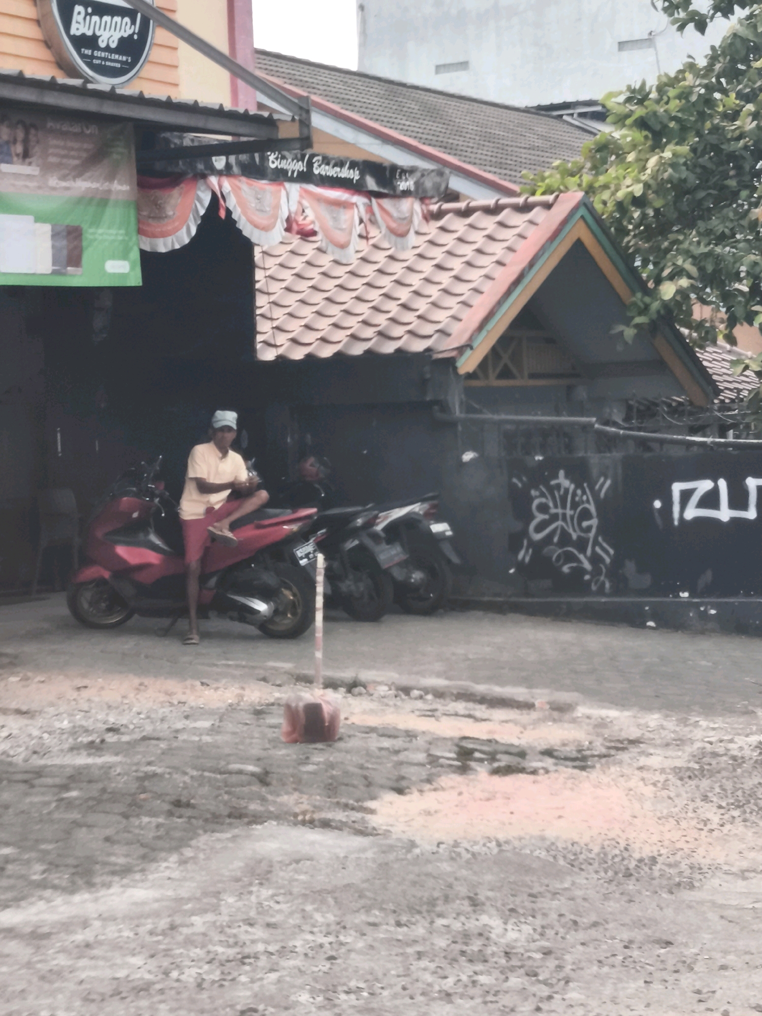 The image captures a street scene, likely in Palembang, Indonesia, with a focus on a barbershop. The barbershop, identified by a sign reading "Binggo! Barbershop," features a prominent orange and pink facade. A banner with the same "Binggo!" logo hangs above the entrance, and decorative orange and white scallops adorn the overhang. In front of the shop, a person wearing a yellow t-shirt and red shorts sits casually on a red scooter. Several other motorcycles are parked nearby. The surrounding environment includes a black wall with white graffiti, trees, and a slightly deteriorated asphalt ground with patches of loose gravel and some pinkish dust. The overall impression is one of a commercial establishment in a typical urban setting.