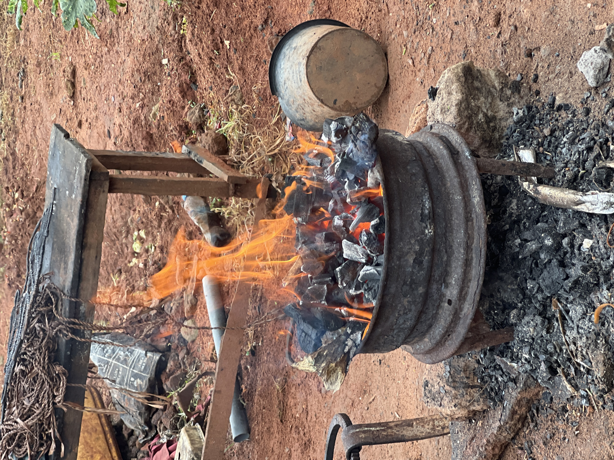 This is a close-up, eye-level shot of an outdoor blacksmithing forge, likely located in the city of Nkpor, Nigeria. The scene is dominated by a large, metal pot filled with glowing hot coals and burning wood, producing vibrant orange flames. The pot is supported by a rusty metal stand, and surrounded by a pile of ash and charcoal. To the left, a portion of a metal tool or implement with a curved handle is visible, possibly a hammer or tongs. In the background, a rough wooden table with a metal top is set up over the fire, with some ropes and a dark, patterned bag hanging from it. A rusty metal pipe lies on the ground behind the fire. To the right, an overturned metal bucket rests on the reddish-brown earth, which makes up the ground surface. The environment appears to be an unpaved area, possibly a yard or workshop, with scattered rocks and sparse vegetation. The overall impression is one of active, traditional craftsmanship.