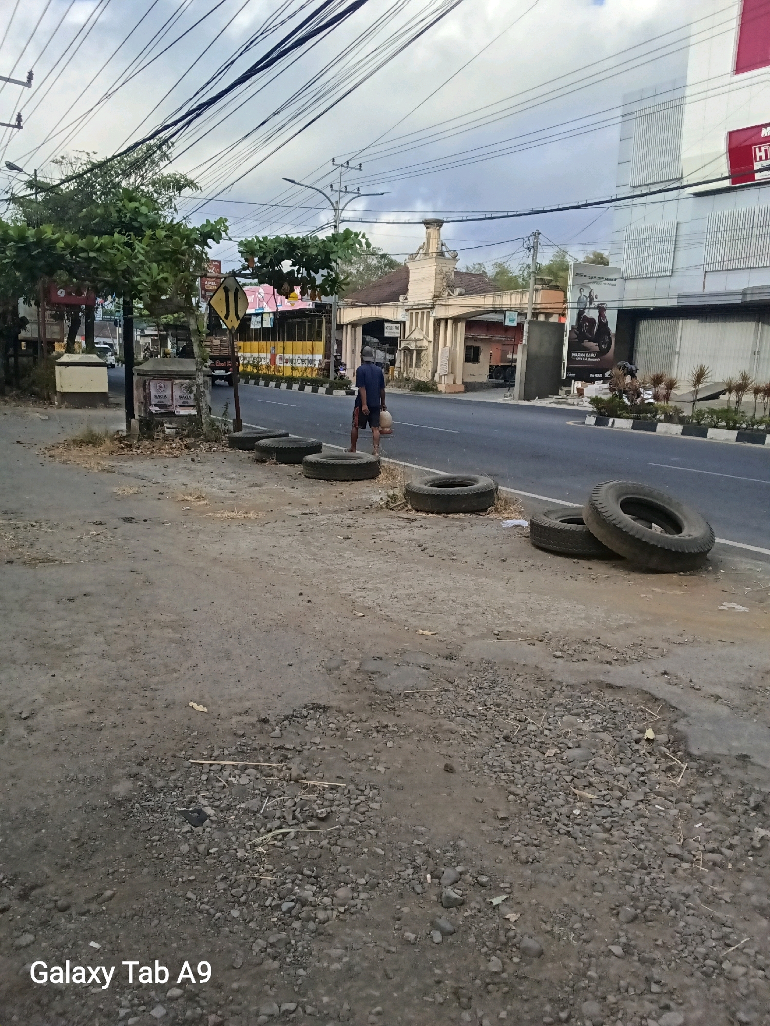 The image depicts a street scene in Trenggalek, Indonesia. A man is walking along the side of the road, carrying a bag. He is wearing a dark blue t-shirt and shorts. Several old tires are scattered on the ground beside the road. In the background, there is a building with a large sign that says "HT". The sky is cloudy. The ground is a mix of dirt and gravel. The area appears to be a commercial or industrial zone.