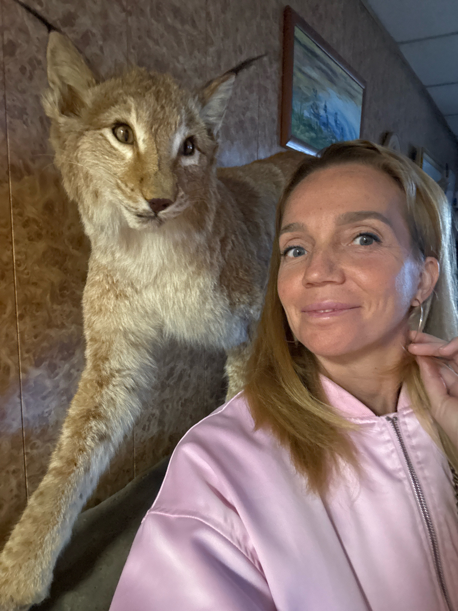 In the city of Успенское, Russia, a woman with blonde hair and blue eyes, wearing a pink bomber jacket, is taking a selfie. She is positioned to the right of a taxidermied lynx, which is mounted on a wall with a wood-patterned wallpaper. The lynx is facing left, with its head slightly turned towards the woman. A framed painting hangs on the wall behind the lynx. The lighting appears to be artificial.