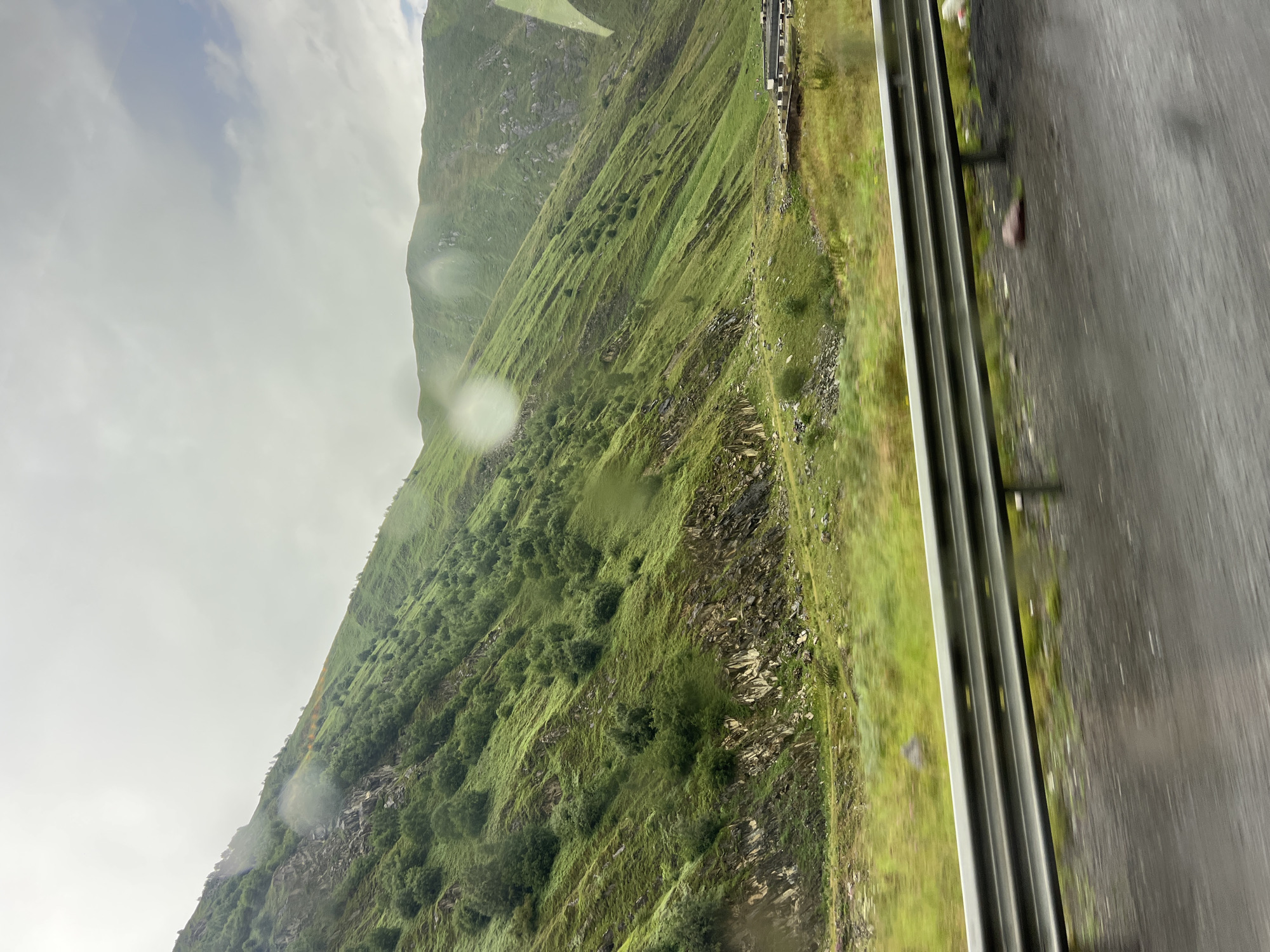 The image depicts a mountainous landscape under a cloudy sky. The mountains are covered in lush green vegetation, with patches of exposed rock. The foreground features a road with a metal guardrail, and the road surface appears wet, suggesting recent rain. In the middle ground, there is a wider, flatter area with more grass and scattered bushes, and a few small structures are visible in the distance. The overall scene conveys a sense of natural beauty and remoteness, characteristic of the mountainous regions of Georgia. The specific location of Almasiani, Georgia is not explicitly identifiable within the image itself without additional context or geographical markers.