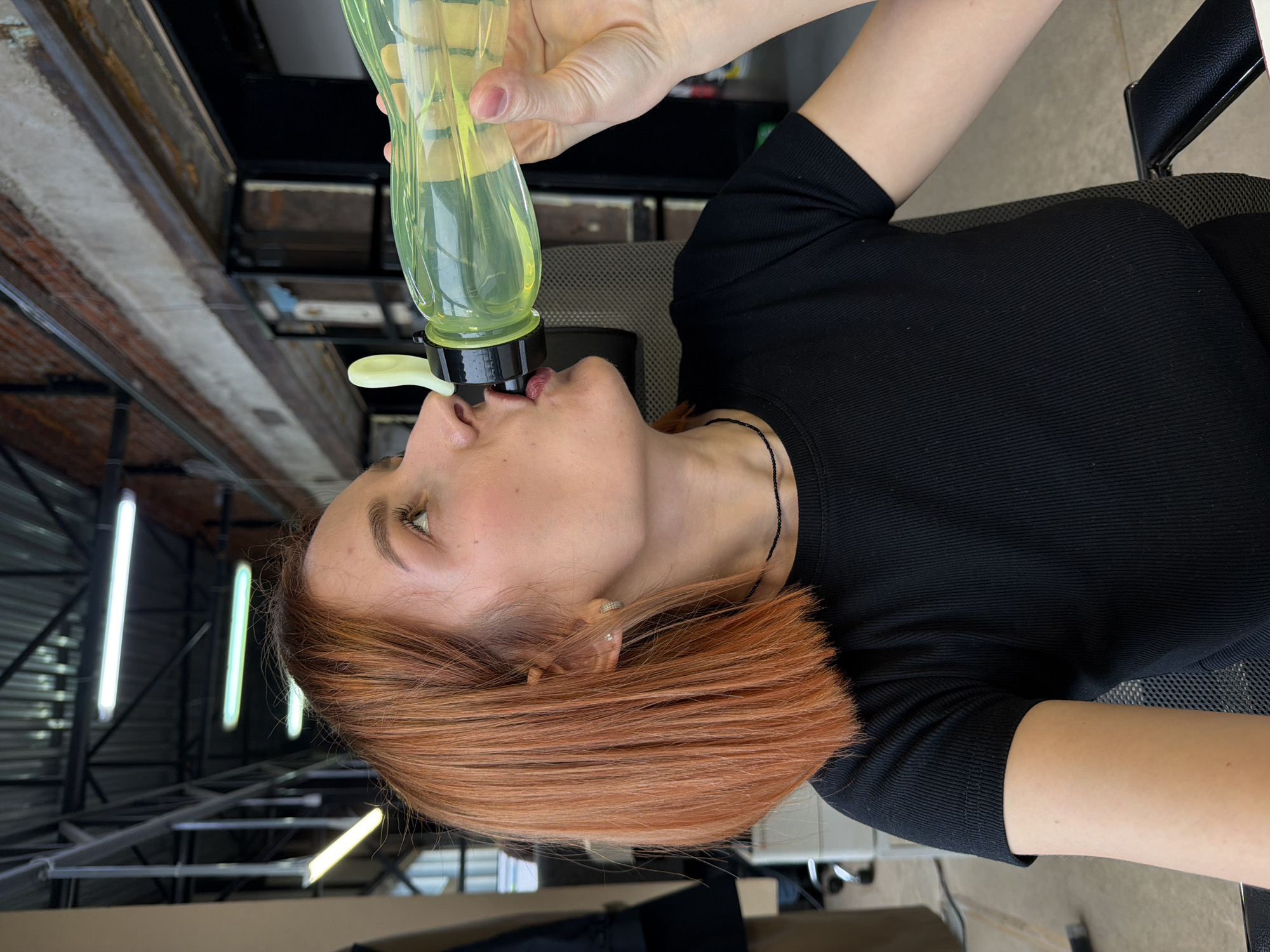 A woman with red hair drinks from a green bottle. The background includes a metal ceiling and a chair. The location is likely inside a building in the city of Chaikovsky, Russia.