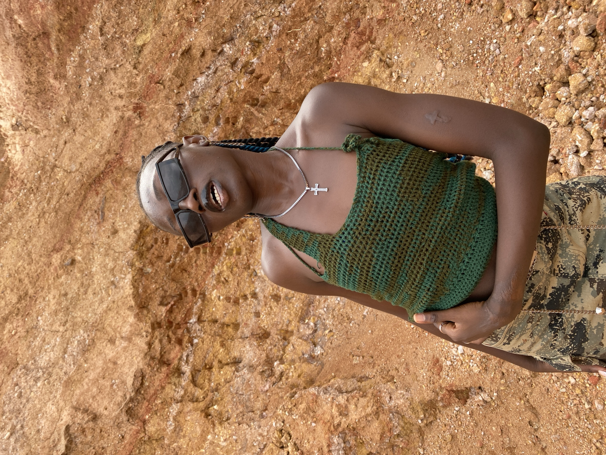 A medium shot captures a young Black woman with dark skin and braided hair adorned with blue streaks. She is positioned against a backdrop of a large, reddish-brown, rocky embankment. The woman is wearing large, black, rectangular sunglasses, a green crocheted halter top, and camouflage-patterned pants. A silver chain with a cross pendant rests on her chest. Her left hand is placed on her abdomen, and a faint cross-shaped marking is visible on her upper left arm. The ground around her is covered with pebbles and rocks. The scene appears to be outdoors in an arid environment, potentially near a construction site or a natural geological formation.