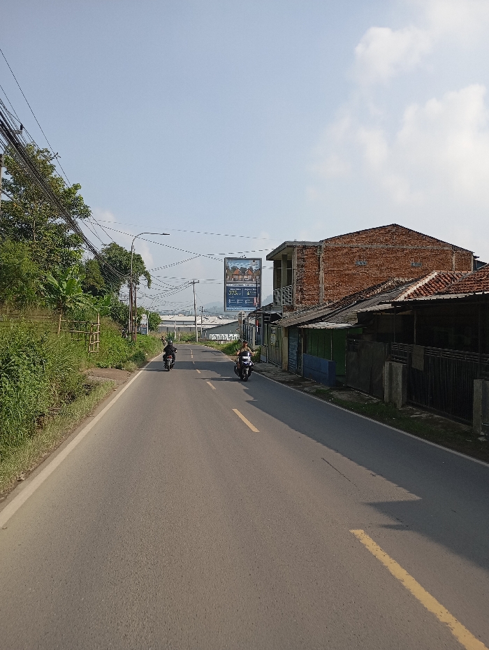 The image depicts a straight asphalt road in the city of Pataruman, Indonesia, observed under a clear blue sky with sparse white clouds. Two motorcycles are visible, traveling away from the viewer in the same direction. One black motorcycle is positioned towards the center-left of the road, further in the distance, with its rider wearing dark attire and a helmet. A second, darker-colored motorcycle is located closer to the viewer on the right side of the road, with its rider wearing a lighter-colored top and a helmet.
On the right side of the road, a series of structures line the street. Prominently, a two-story building with exposed red brick walls and a dark, possibly unfinished, upper section is visible, featuring a tiled roof. Adjacent to it are various single-story buildings with corrugated metal roofs and walls of different colors, including blue, green, and grey, fronted by a dark metal fence or gate.
The left side of the road is bordered by an elevated embankment covered in dense green vegetation, including bushes and banana-like plants. Multiple utility poles rise from this side, supporting numerous tangled black electrical and communication wires that stretch across the road. A street light is affixed to one of these poles.
In the mid-ground, a large billboard advertising "GOLDEN HILLS" residential properties with pricing ("375jt") and payment terms ("Angsuran Ringan") stands on the right. Distant hills or mountains are visible on the horizon, beneath a clear sky. Shadows from unseen objects extend from the left side onto the road, indicating a high sun position.
