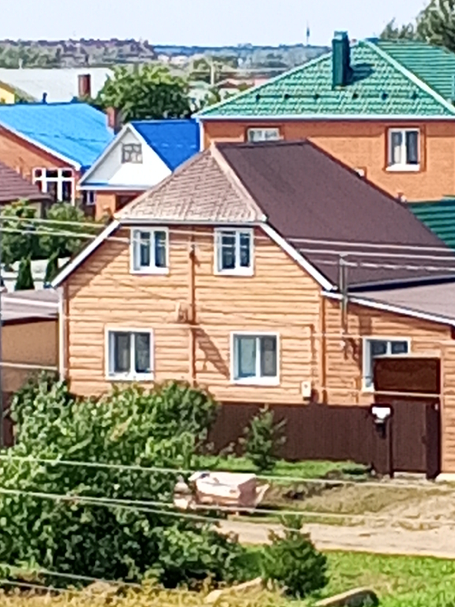 The image shows a cluster of residential buildings in Chistopol, Russia. The foreground features lush green foliage and a dirt path. In the mid-ground, a prominent wooden house with a brown roof and visible log construction is centered. It has several white-framed windows. Adjacent to this house is a smaller building with a light brown roof and white trim. Behind these structures, various other houses are visible, including one with a bright blue roof and another with a green roof. The background reveals a hazy landscape with trees and distant structures, suggesting a suburban or semi-rural setting within Chistopol. There are no people or discernible actions depicted in the scene.