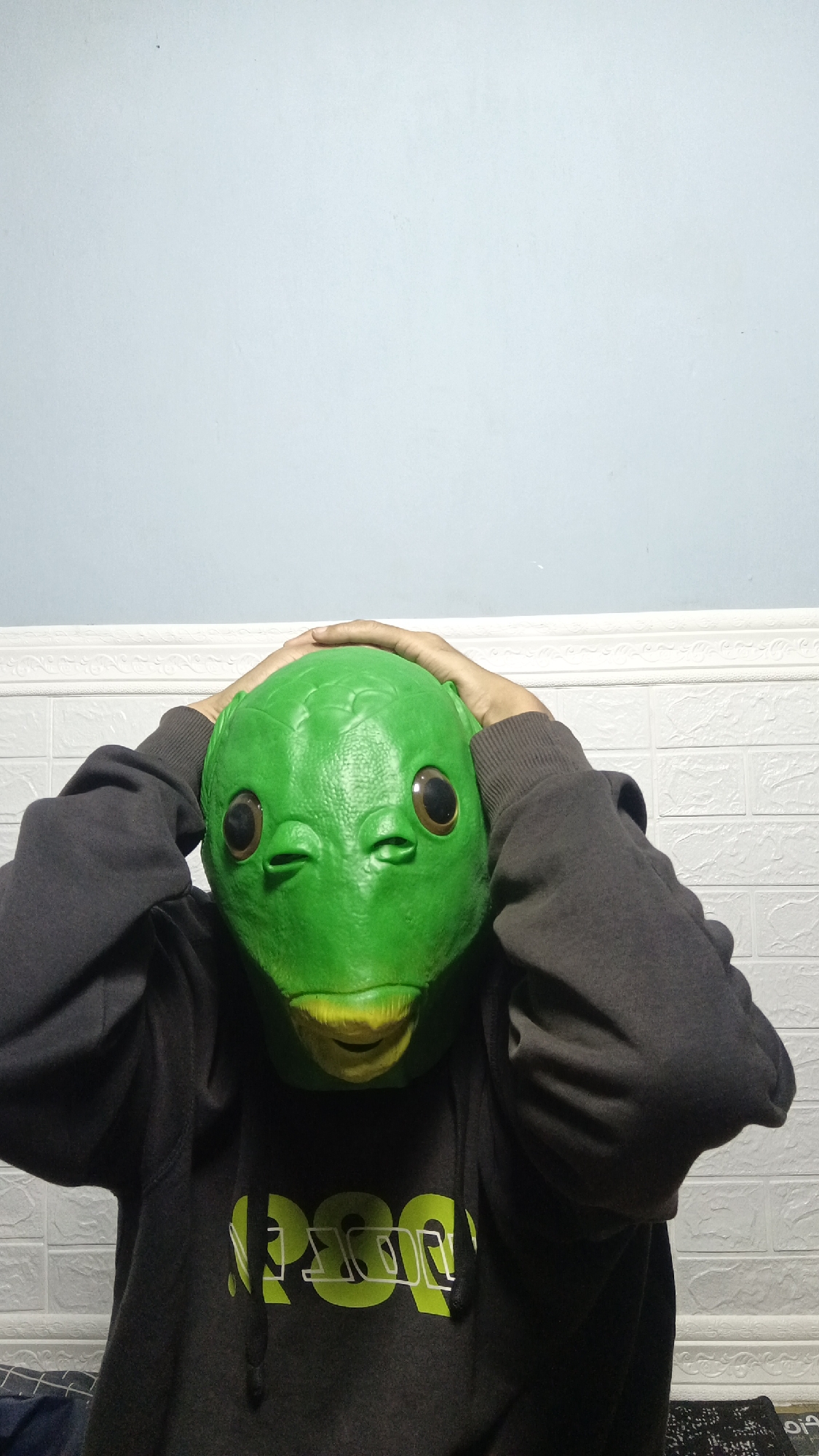 A person wearing a green alien mask and a dark grey hoodie stands in a room. The person's hands are on top of the mask, covering their ears. The background shows a white tiled wall with a light blue upper wall. The hoodie has a green graphic design on the front. The image was taken in the city of Kebumen, Indonesia.