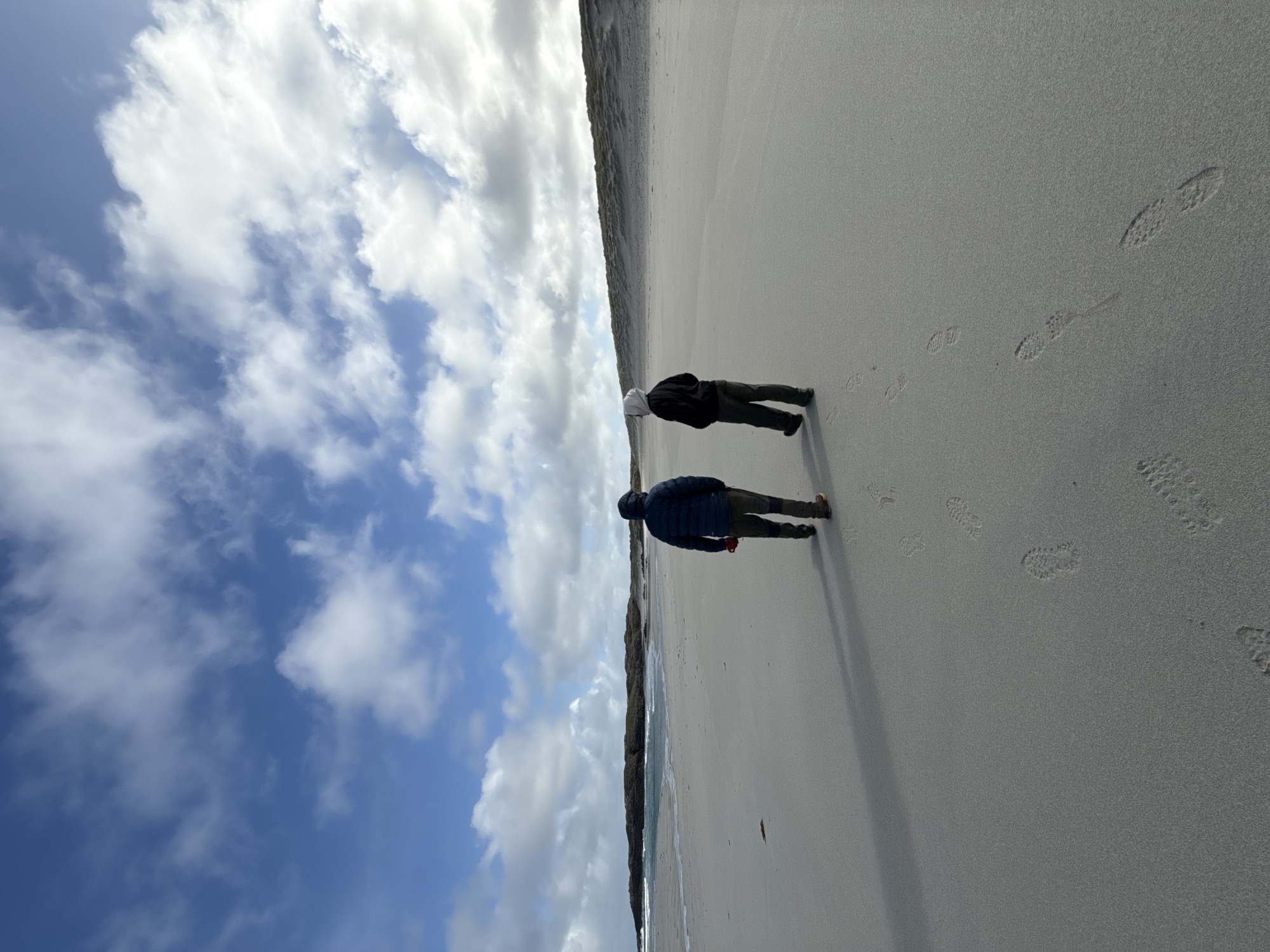 Two people walk on a sandy beach under a partly cloudy sky. Footprints are visible in the sand. A vegetation line borders the beach. The location is the city of Unknown, Unknown.