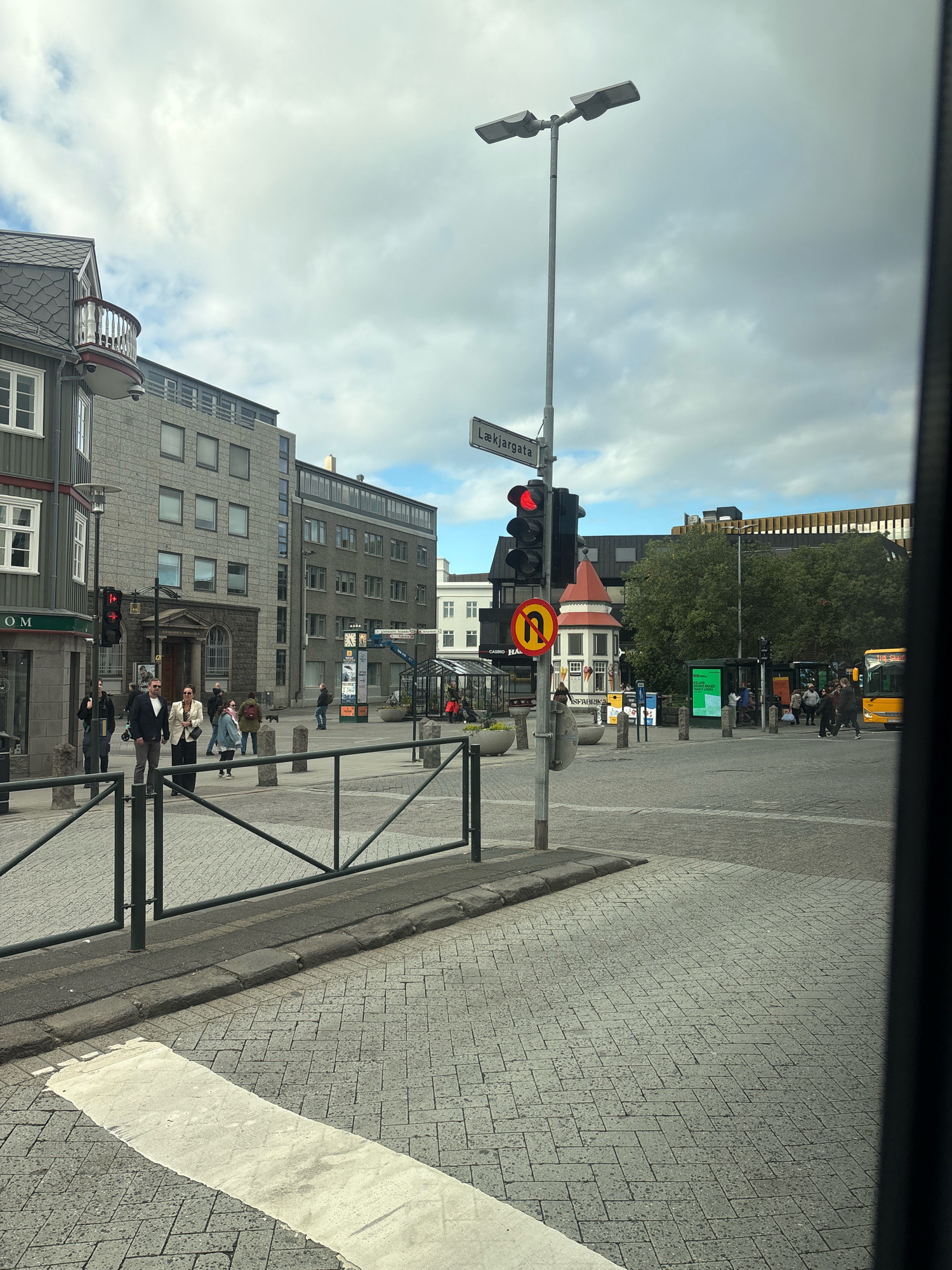 The image shows a street scene in Reykjavik, Iceland, focusing on an intersection with a traffic light and street signs. A traffic light is red, and a "no U-turn" sign is visible. Several buildings line the street, including a modern grey building with multiple windows and a building with a dark grey tiled roof and a balcony. People are walking on the sidewalk and crossing the street. A yellow bus is parked at a bus stop. The ground is paved with grey cobblestones. The sky is cloudy. The scene appears to be a typical urban environment in Iceland.