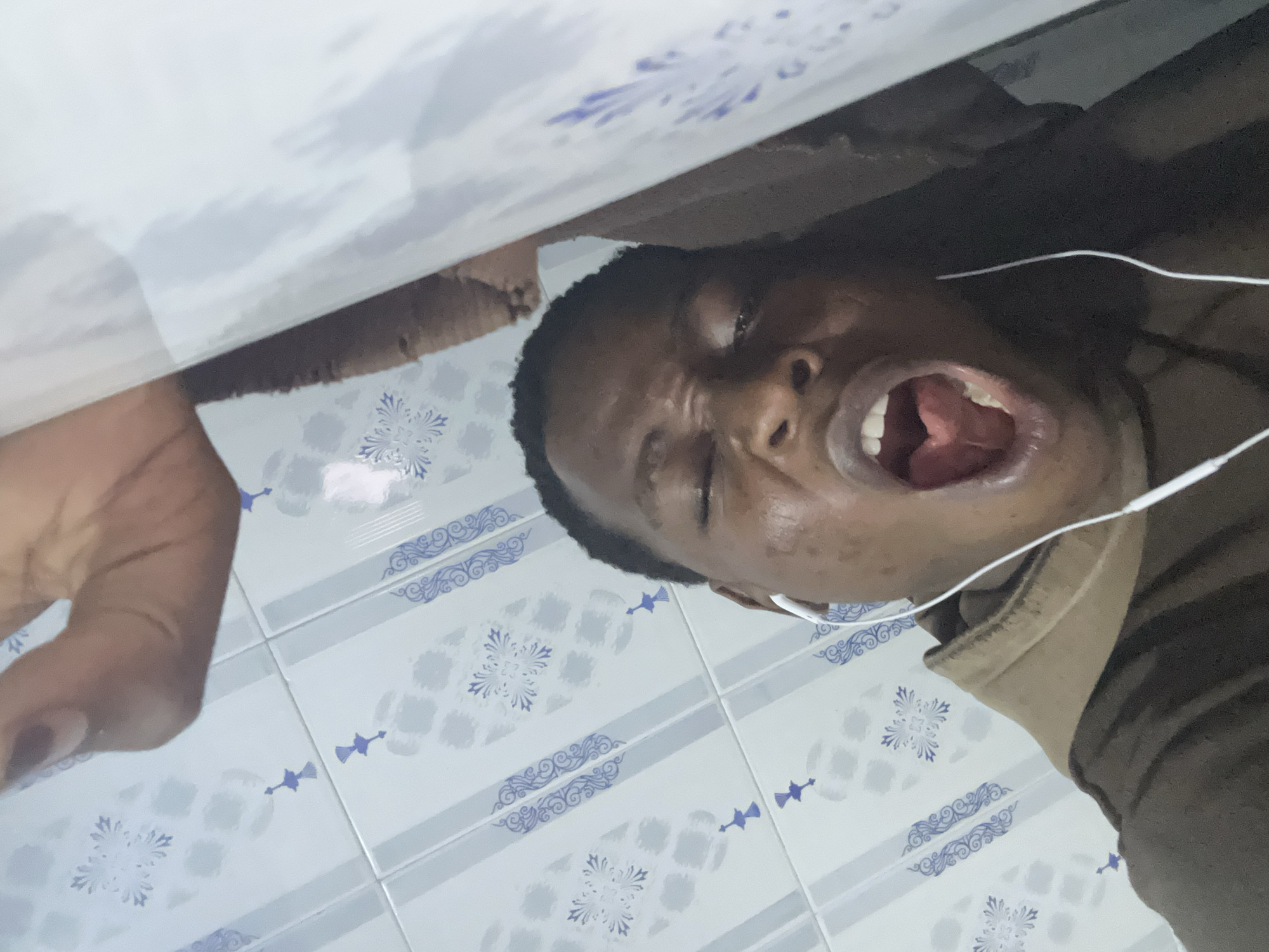 A person with dark skin and short black hair is yelling. The person is wearing earphones. The background consists of white and blue patterned tiles and a white and beige wall. The location is Unknown, Unknown.