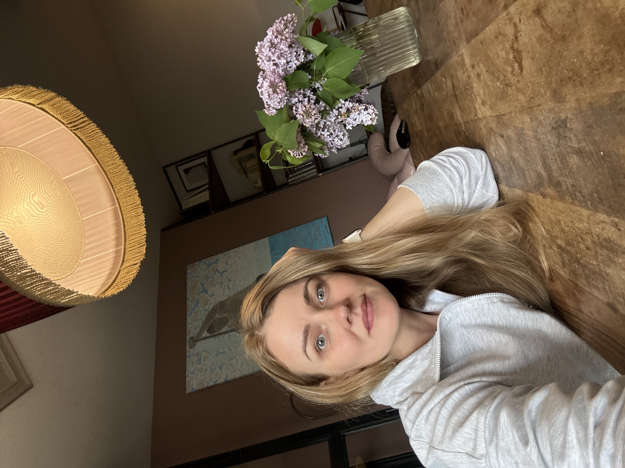 The image shows a woman with blonde hair wearing a gray hooded sweatshirt. She is leaning on a wooden table. A vase of lilacs is on the table near her. Behind her are a painting of a bird, some framed items, and a wall painted a light brown. A lamp with a fringed shade hangs in the corner of the room. The image does not provide enough information to determine if the location is in Saint Petersburg, Russia.