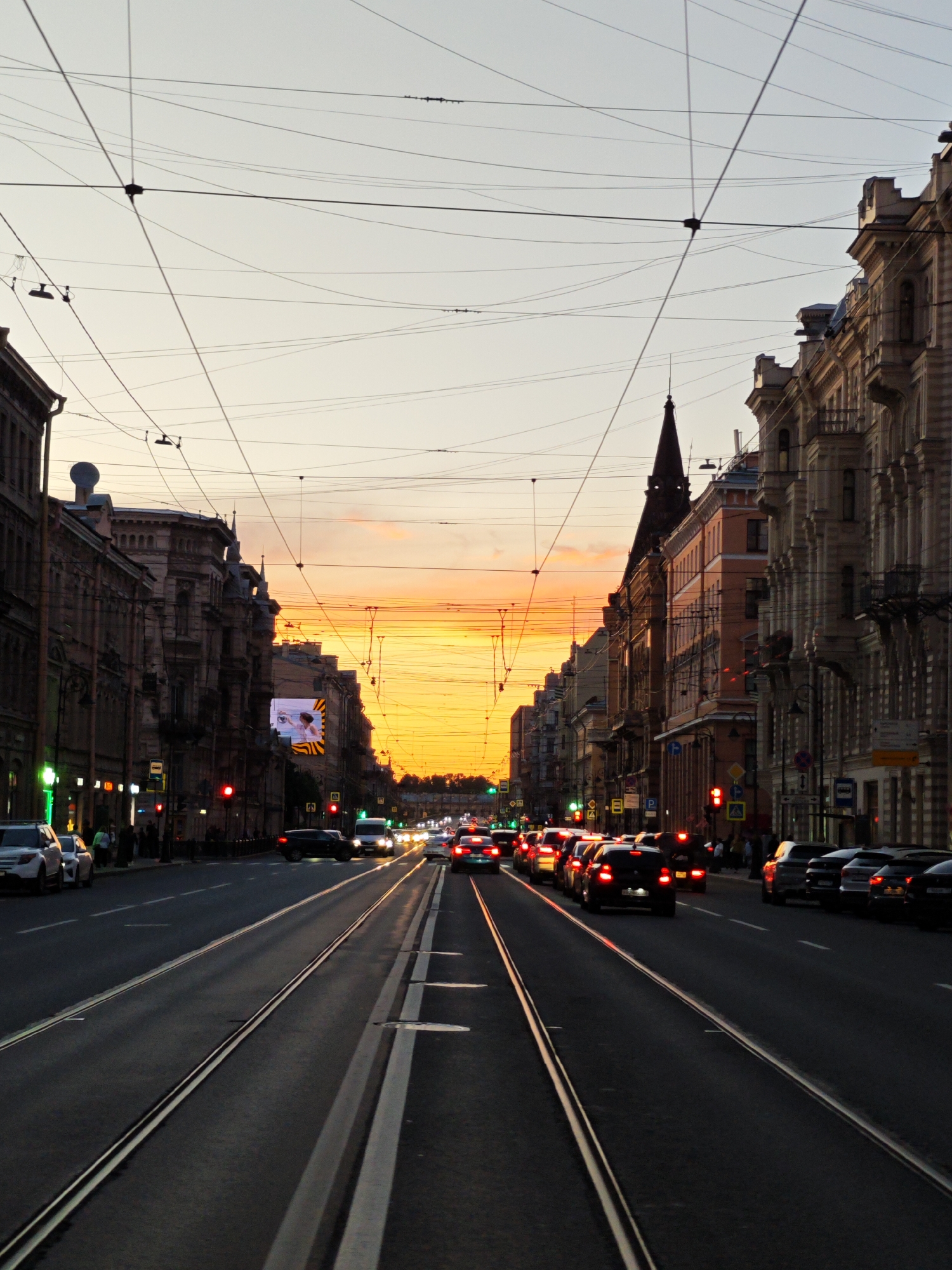The image depicts a street scene in Saint Petersburg, Russia, during twilight. The sky is a gradient of pale yellow to light orange, indicating sunset or sunrise. Numerous overhead wires, likely for trams or trolleybuses, crisscross the frame, creating a complex network against the sky.
On both sides of the street are multi-story buildings with ornate architectural details, typical of Saint Petersburg's historical center. Some buildings have decorative facades, balconies, and classical elements. The street itself is paved, with tram tracks running down the center.
A considerable amount of traffic is present, with cars and possibly buses moving along the road. Red traffic lights are visible, suggesting a busy intersection or congestion. There are also pedestrians on the sidewalks, though they are not the main focus of the image. A digital billboard is mounted on one of the buildings.
The overall atmosphere is urban and dynamic, with the warm colors of the sky contrasting with the darker tones of the buildings and the street. The presence of tram tracks, historical architecture, and the specific lighting conditions strongly suggest a location within the central historical district of Saint Petersburg.