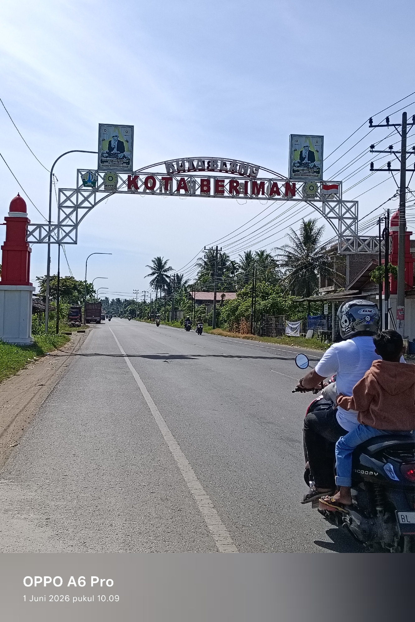 The image depicts a road scene in Tingkeummanyang, Indonesia. A black scooter with two occupants is in the foreground. The driver, wearing a white shirt, dark pants, a helmet, and sandals, is operating the scooter. A child in a brown jacket and jeans sits behind him.
In the middle ground, the road extends into the distance with a white line marking the center. Several other motorbikes are visible further down the road.
An ornate archway spans the road. The archway is decorated with the words "KOTA BERIMAN" and "KULABLANG." On either side of the road are red and white pillars. Trees and vegetation line the road. Utility poles and wires are visible. The sky is light blue with some clouds.