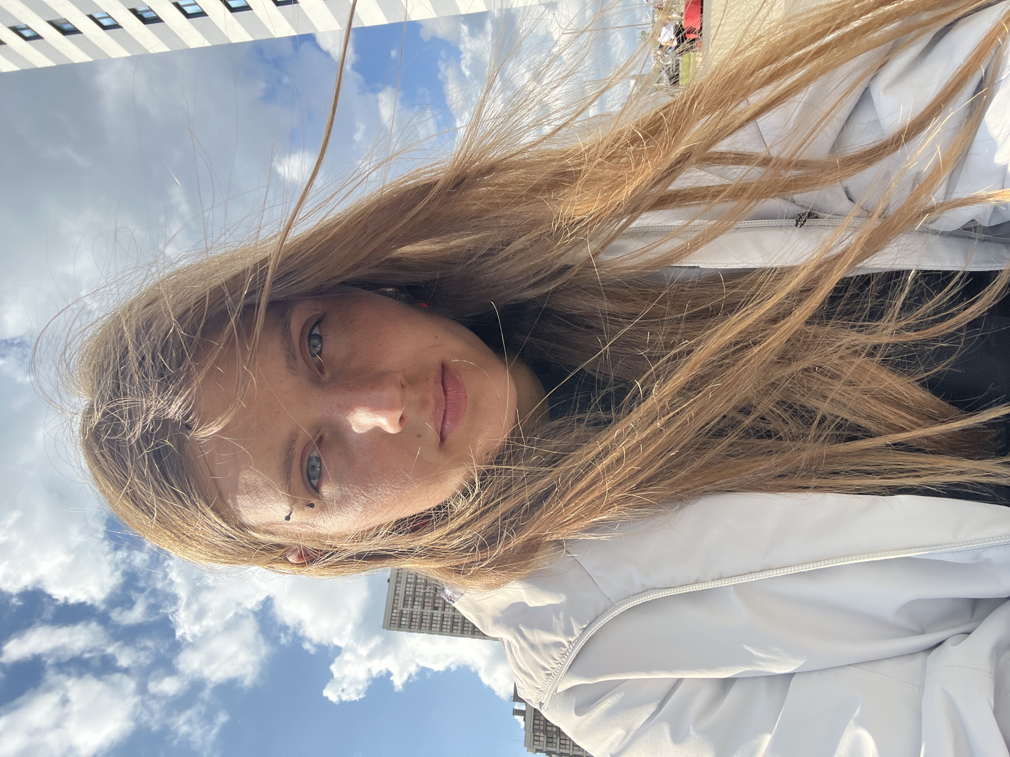 A selfie taken from a low angle shows a young woman with blonde hair and freckles looking directly at the camera. She is wearing a white jacket over a dark garment. She has a piercing above her left eyebrow. Her hair is blowing in the wind. Behind her, a bright blue sky with fluffy white clouds is visible, along with the upper floors of modern apartment buildings. The overall impression is one of an outdoor urban setting on a sunny day.