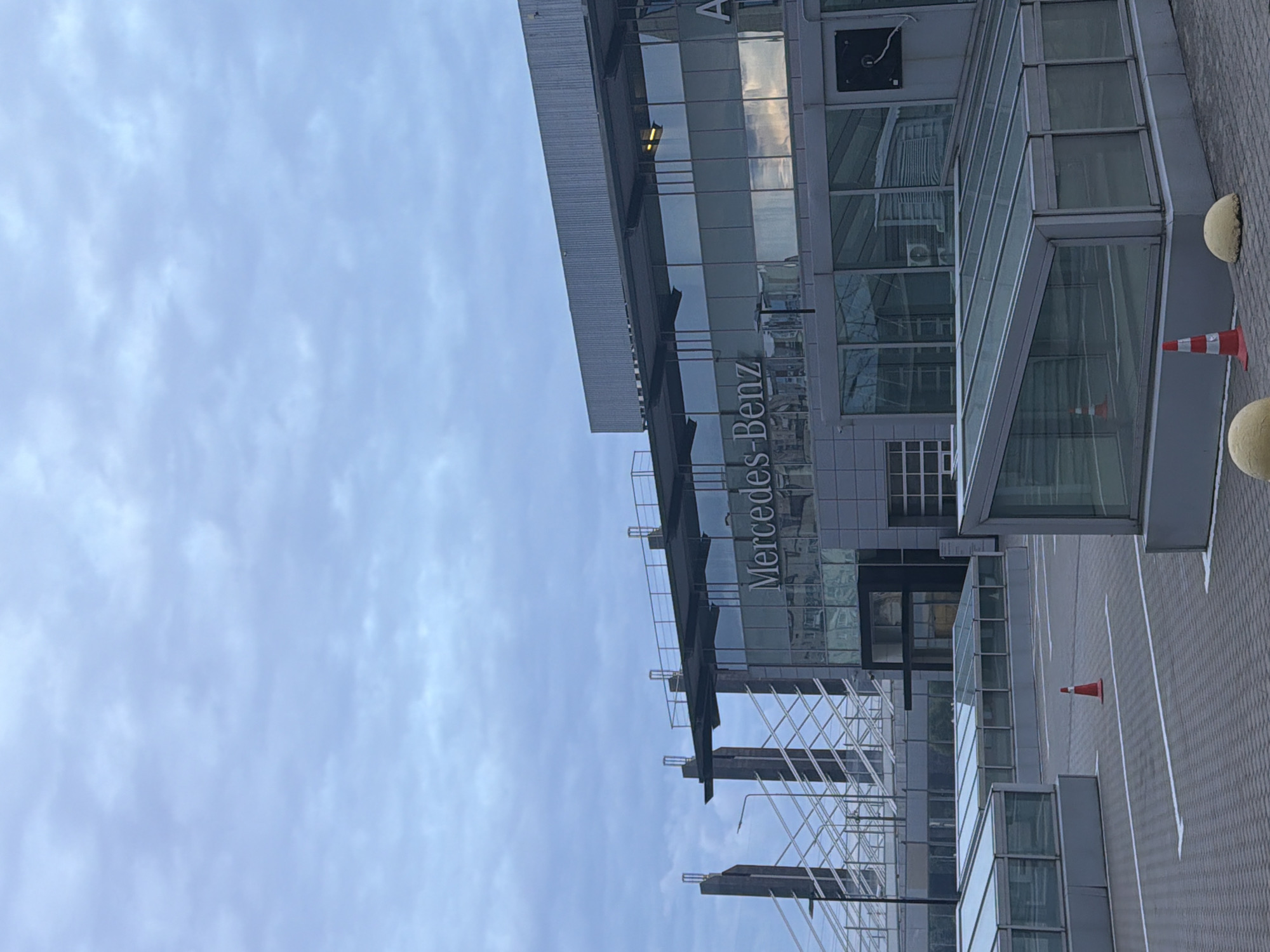 A Mercedes-Benz dealership is visible under a cloudy sky. The building has a modern, angular design with large glass windows and a metal facade. The "Mercedes-Benz" logo is prominently displayed on the upper level of the building. In the foreground, there is a paved area that appears to be a parking lot or showroom, with several glass-enclosed structures and orange traffic cones. In the background, there are tall, structural elements that resemble scaffolding or part of a larger infrastructure project. The overall impression is that of a commercial building in an urban setting.