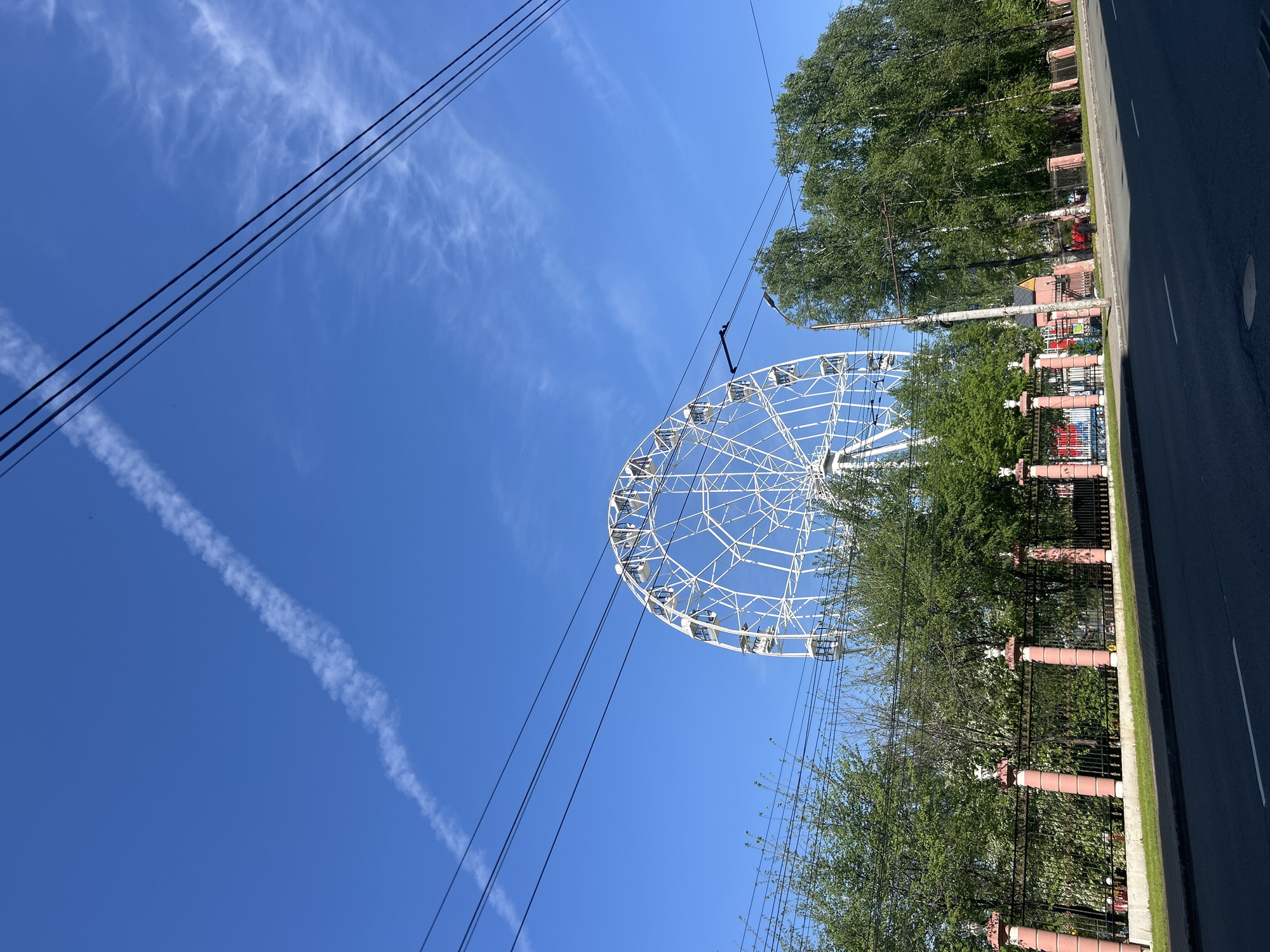 The image is taken from a low angle looking upwards, showing a ferris wheel partially obscured by trees, set against a clear blue sky. The ferris wheel is white and has several gondolas visible. Power lines extend across the top left portion of the frame. The side of a road and a decorative fence line the right edge of the frame, separating the road from the trees partially blocking the wheel. A contrail is visible in the sky near the upper-left quadrant. Based on the characteristics of the landmarks depicted in this image, this photo was likely taken in Izhevsk, Russia.