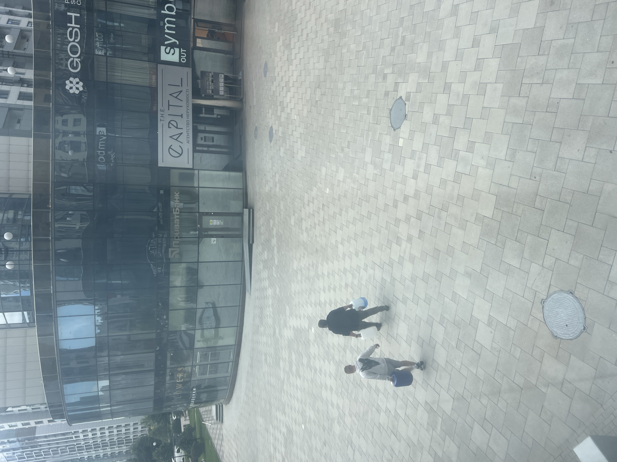 This is an exterior, eye-level shot of a modern commercial building in Kyiv, Ukraine, on an overcast day. The building features a curved glass facade with multiple storefronts. Visible signage includes "GOSH", "THE CAPITAL", and "Symbo" in English, along with Ukrainian text such as "ПУМБ" and "АГЕНТСТВО НЕРУХОМОСТІ". Two men are walking across a wide, paved plaza in front of the building. The man on the left is wearing a grey hooded sweatshirt and shorts, carrying a large blue container. The man on the right is wearing a dark sweatshirt and dark pants, carrying a white and a blue container. The plaza is paved with light-colored square tiles. Several circular manhole covers are visible on the ground. Tall apartment buildings are visible in the background to the left of the commercial structure.