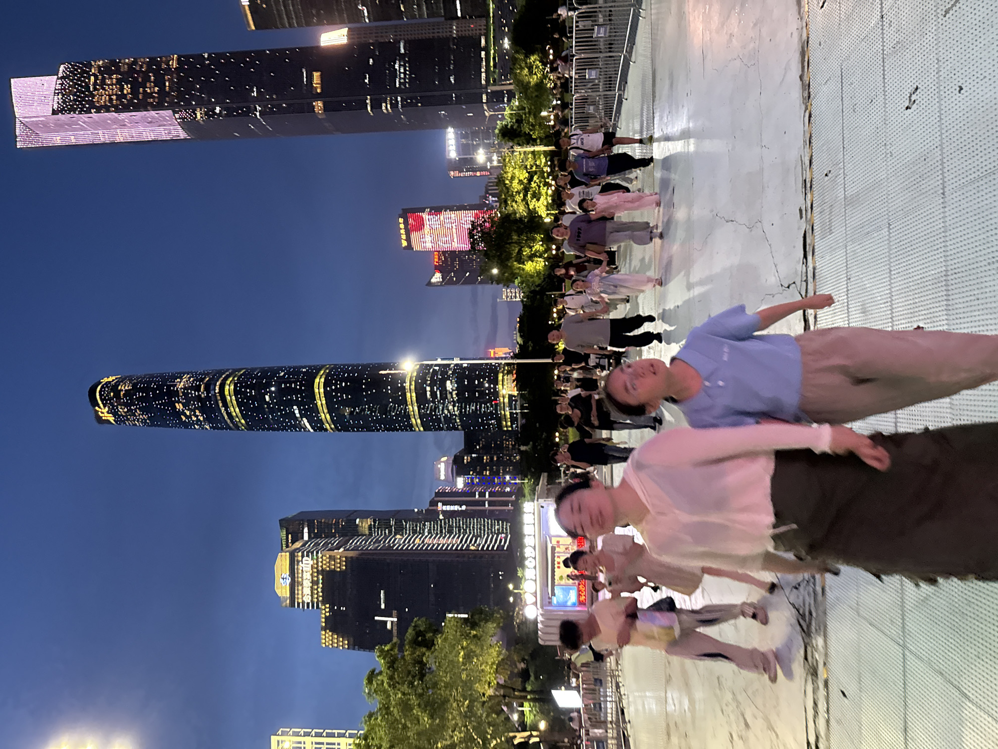 The image depicts a nighttime scene in the Tianhe District of Guangzhou, China, likely in the vicinity of the Guangzhou International Finance Center and the CTF Finance Centre, both prominent skyscrapers visible in the background. A crowd of people is walking across a paved outdoor area, illuminated by the ambient city lights and the glow of the surrounding buildings.
In the foreground, two women are walking towards the viewer. The woman on the left is wearing a white open cardigan over a darker garment and a long, dark skirt. The woman on the right is wearing a light blue polo shirt and light-colored pants. Several other individuals and groups are visible further back, including a family walking hand-in-hand, men and women dressed casually, and children. The architecture of the city is a dominant feature, with tall, modern skyscrapers lit up with patterns of lights, creating a visually striking urban landscape.
A small kiosk or stand is situated in the mid-ground, with text and images displayed on its facade. Metal barriers line the right side of the paved area. The sky is a deep blue, indicating it is early evening or dusk. The overall impression is of a busy, well-lit urban environment at night, with people engaged in evening strolls or activities.