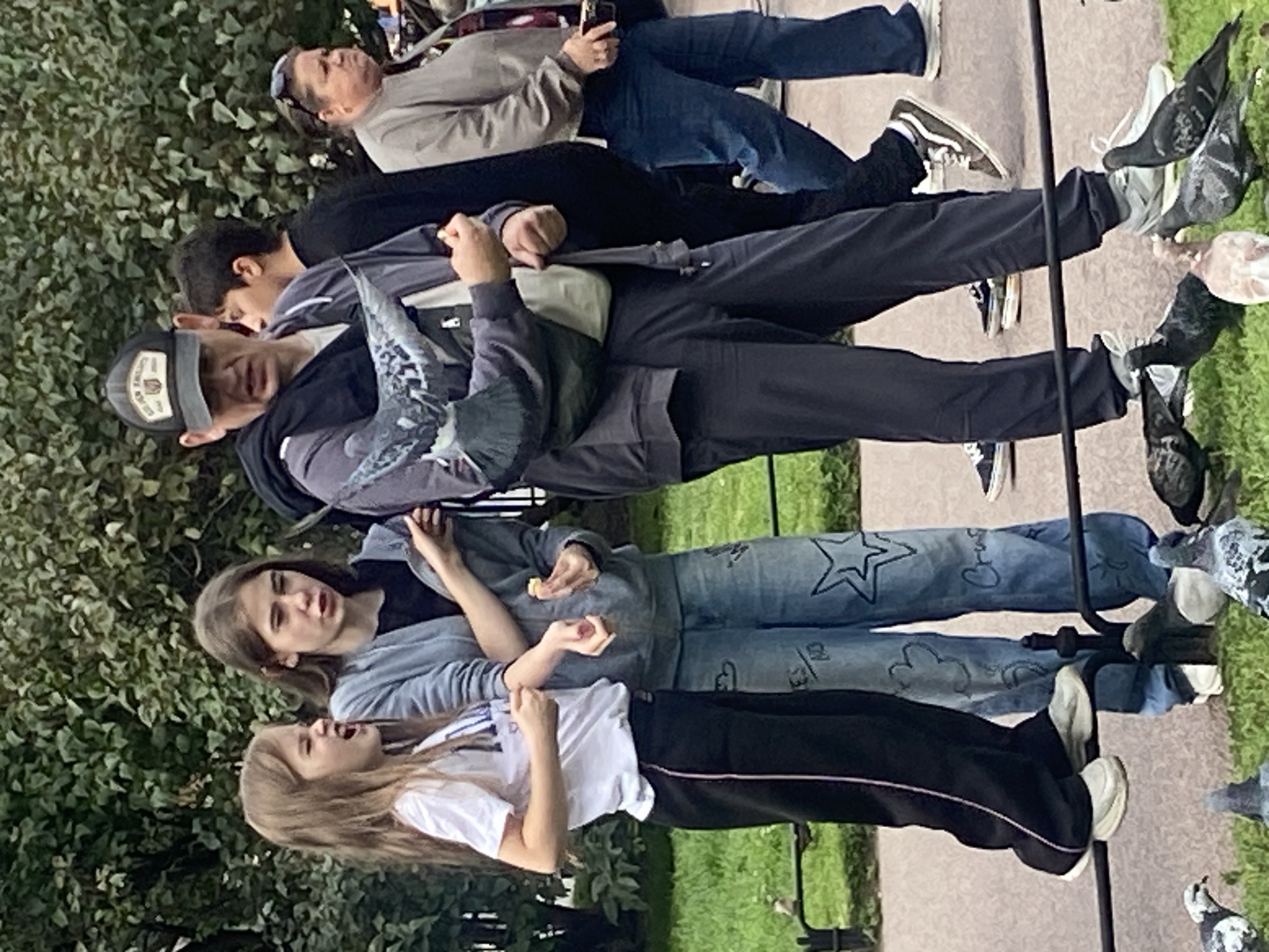 In Saint Petersburg, Russia, a group of people are in a park with trees and grass. A man in a baseball cap holds a pigeon on his arm. Two young girls are standing next to him, one with her mouth open as if speaking or yelling. The other girl is holding a piece of food. Several pigeons are on the grass in the foreground. Other individuals are visible in the background.