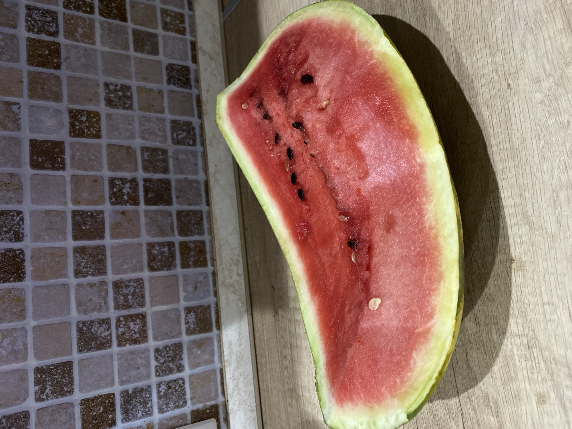 A slice of watermelon with seeds is positioned on a wooden surface in front of a tiled backsplash. The watermelon is brightly colored with a pinkish-red flesh and a green rind. The backsplash features small, square tiles in varying shades of brown and beige, arranged in a grid pattern. The lighting suggests an indoor setting, possibly a kitchen. The scene is static, with no visible people or activity. The image does not provide any information about its location in Chișinău, Moldova.